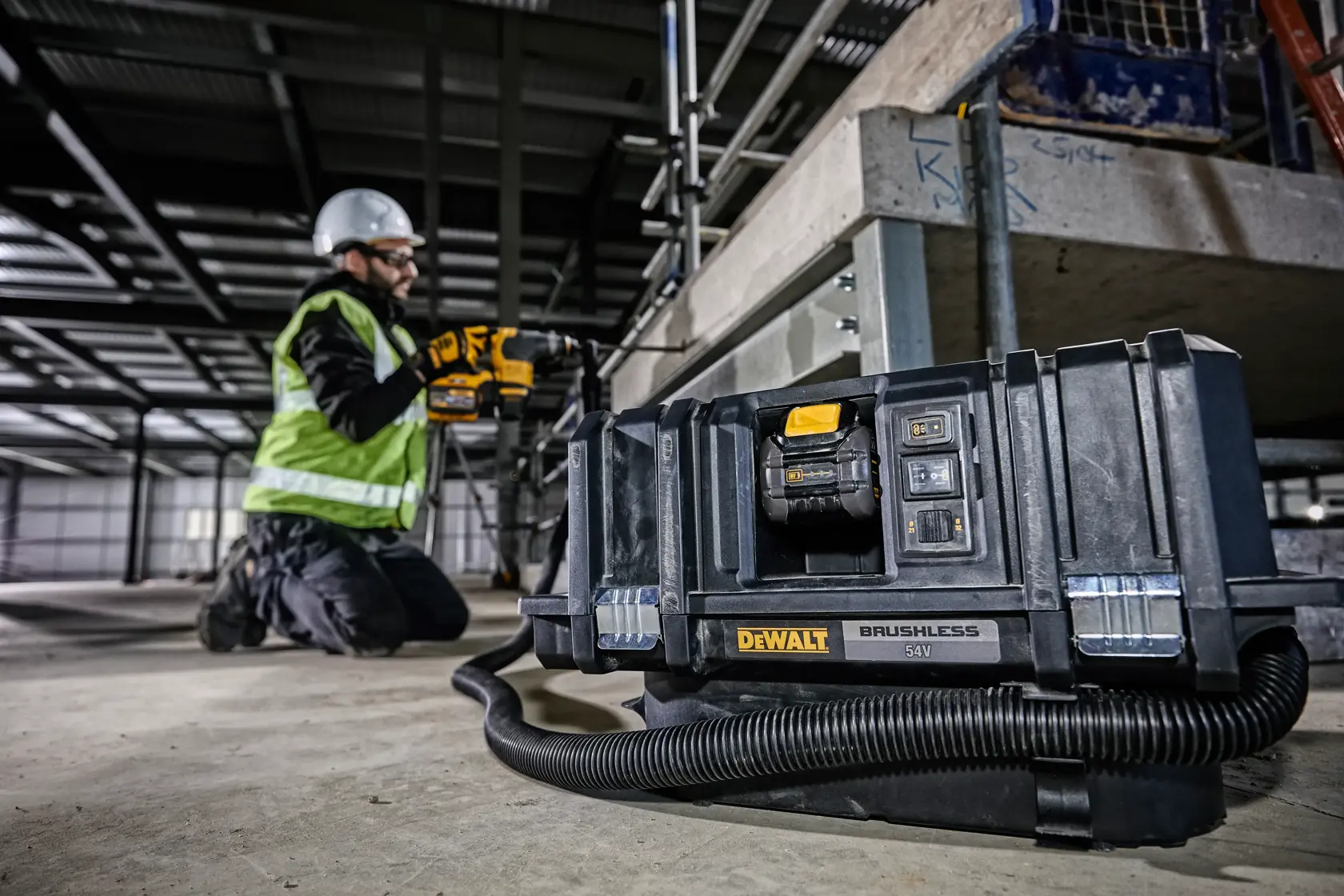 DEWALT DCV586MT2 brushless vacuum on a construction site floor, worker kneeling nearby in safety gear.