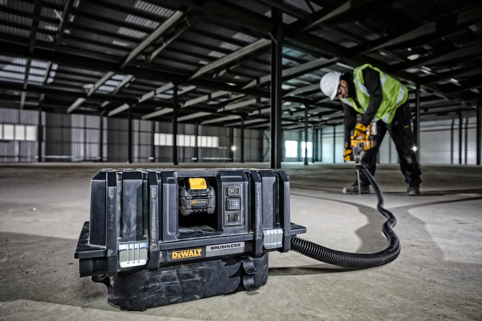 DEWALT DCV586MT2 vacuum on construction site floor, worker drilling in background wearing safety gear.