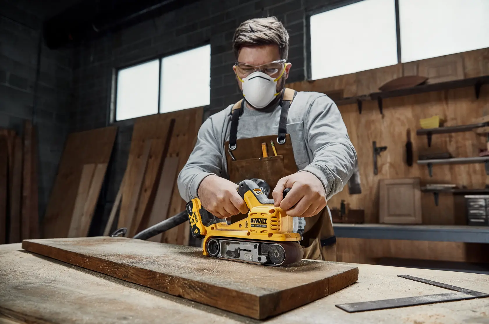 En person bruker en gul DEWALT DCW220P2 trådløs beltessliper til å slipe et trappetrinn i tre. En støvsugerslange er koblet til verktøyet for støvoppsamling.