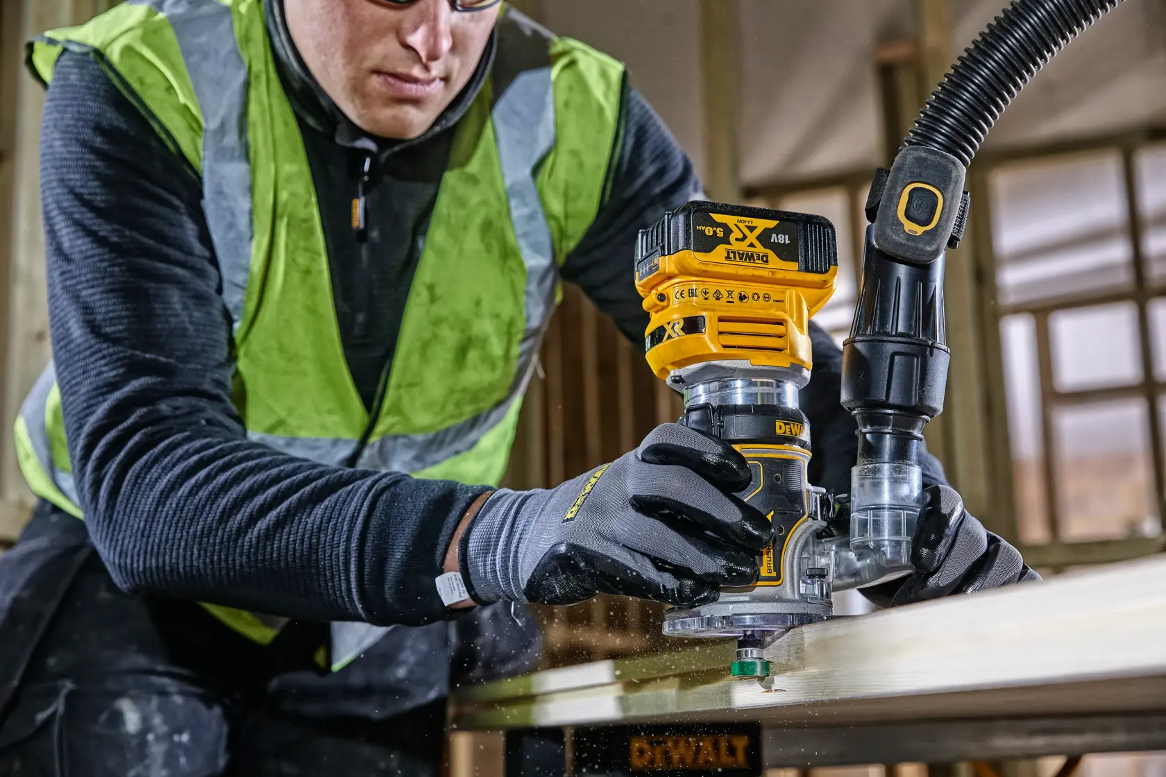 Trabajador usa una fresadora DEWALT DCW604 en madera, con guantes y chaleco de seguridad.