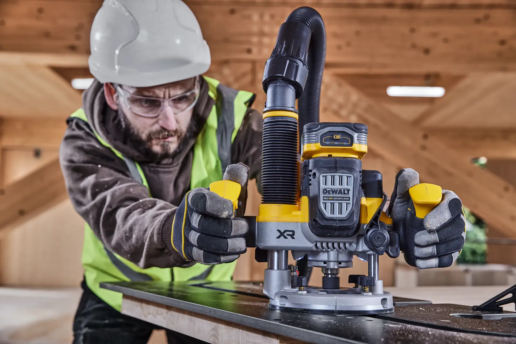 Worker using DEWALT XR DCW620H2 cordless router on wood in construction site, wearing safety gear.