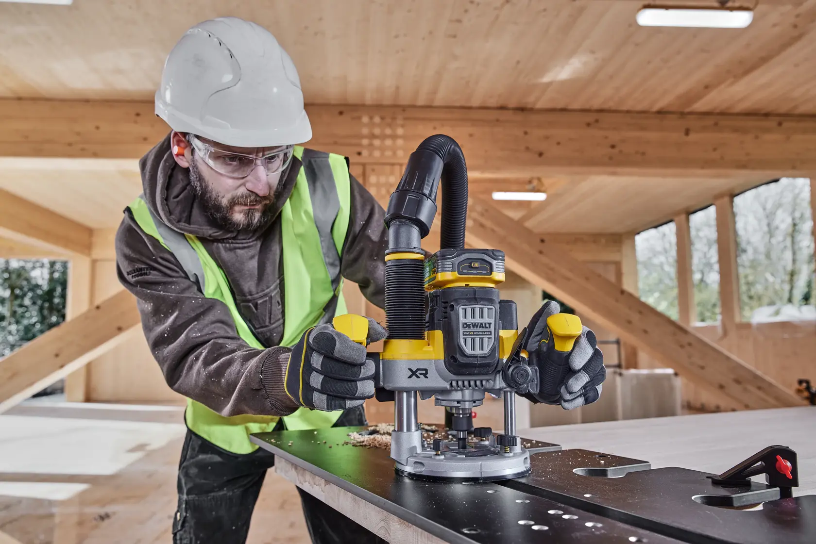 Worker uses DEWALT XR router DCW620H2 on a board in a wood structure, wearing safety gear.
