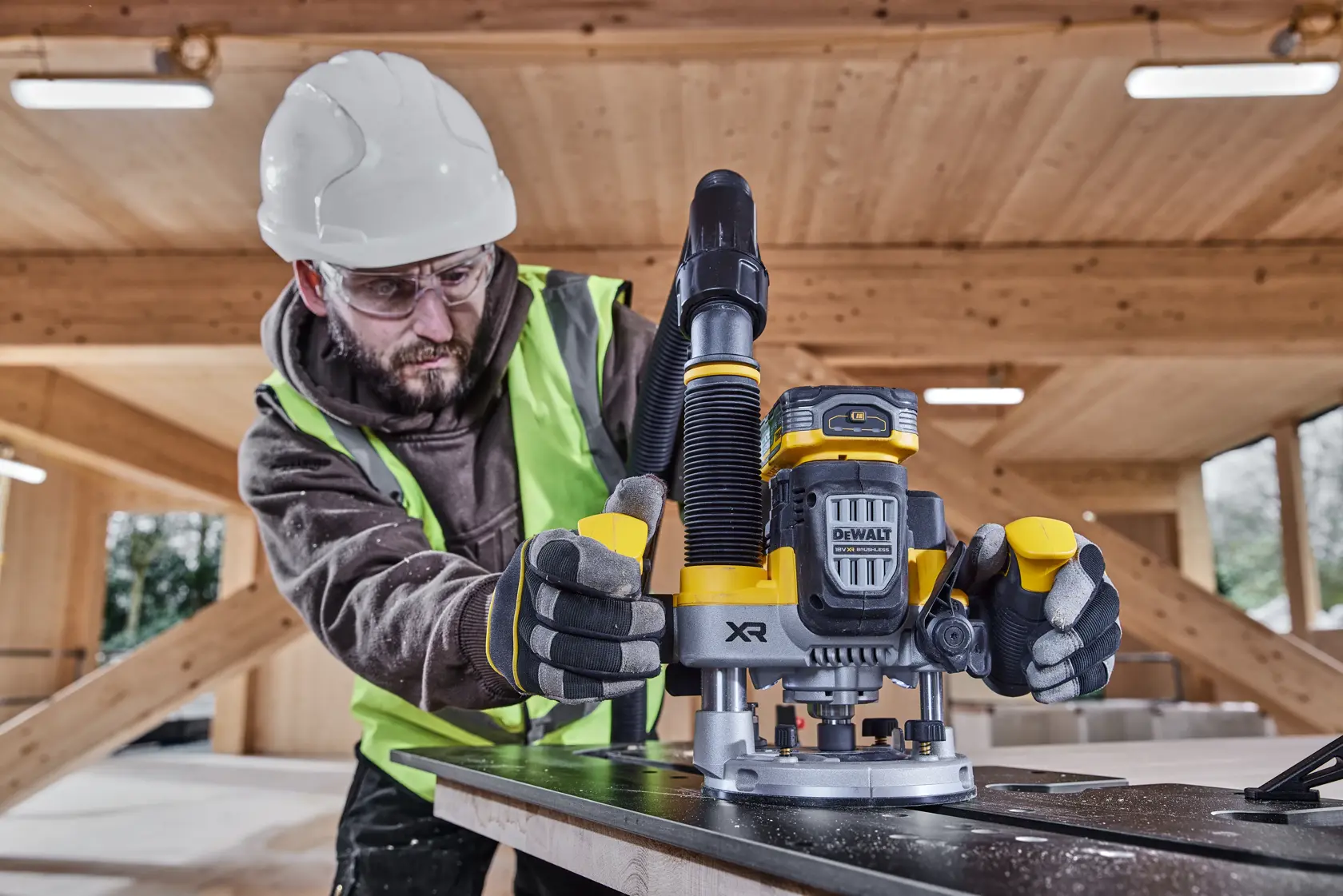 Worker uses DEWALT cordless router DCW620H2 on wood in a construction site, wearing safety gear and gloves.