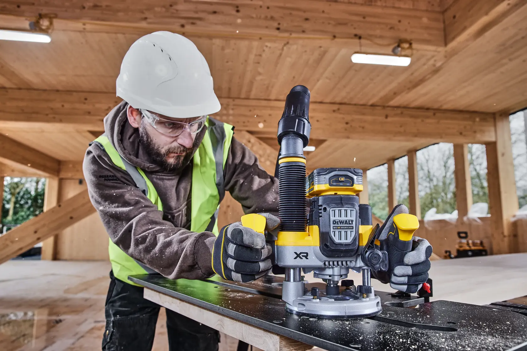 Worker uses a DEWALT DCW620H2 router on wood in a construction setting, wearing safety gear and gloves.