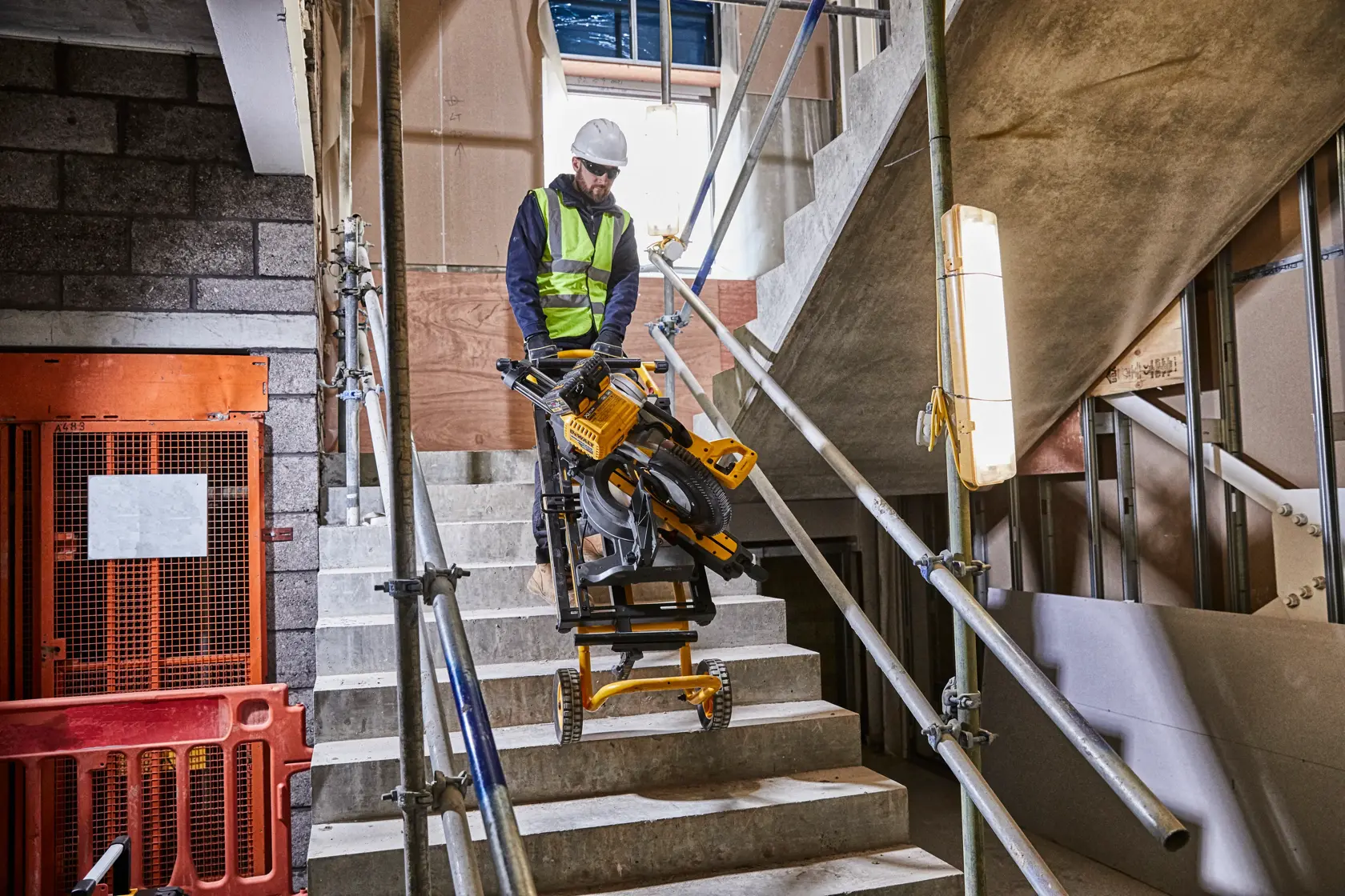 Worker moves DEWALT saw and stand up stairs at a construction site with safety rails and barriers visible.