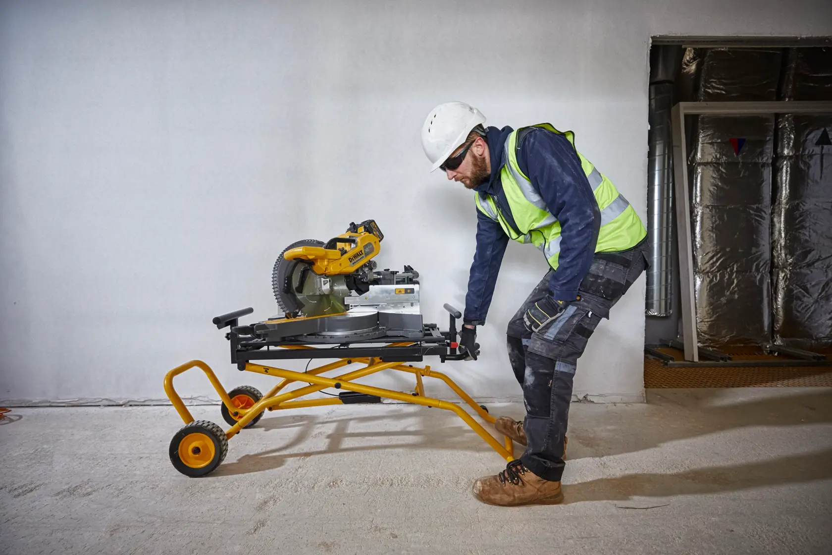 Worker moves a DEWALT miter saw on a yellow wheeled stand inside a construction site.