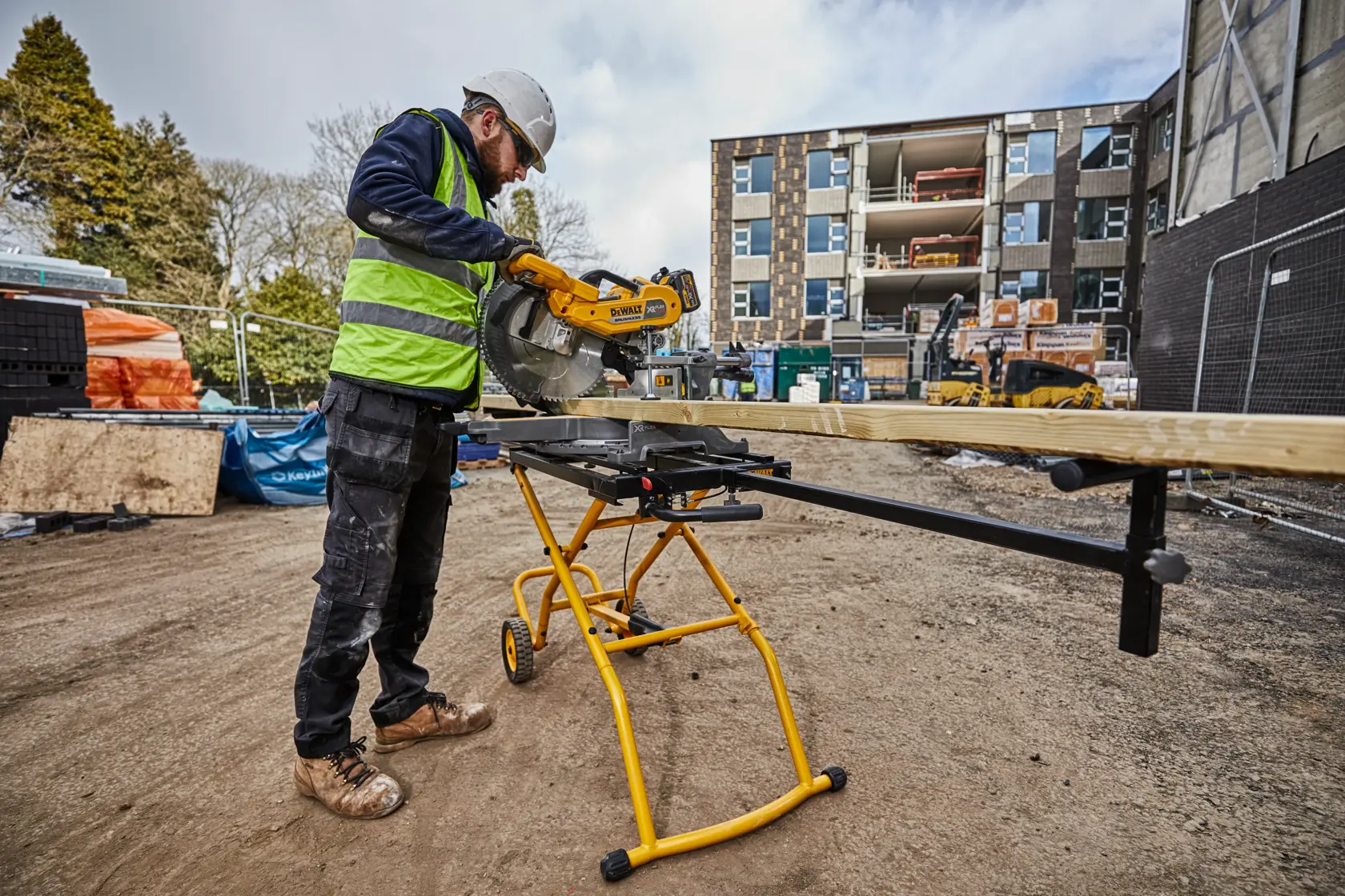 Worker uses DEWALT saw stand DE7260 to cut wood at a construction site. Building and equipment visible in background.