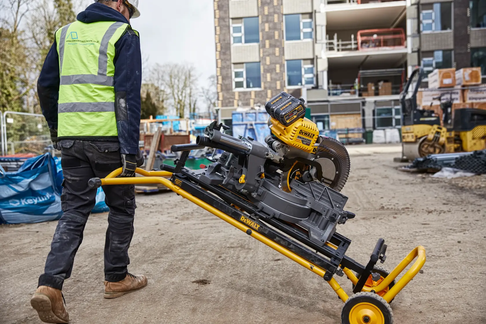 Worker moves a DEWALT miter saw on a rolling stand at a construction site with buildings in the background.