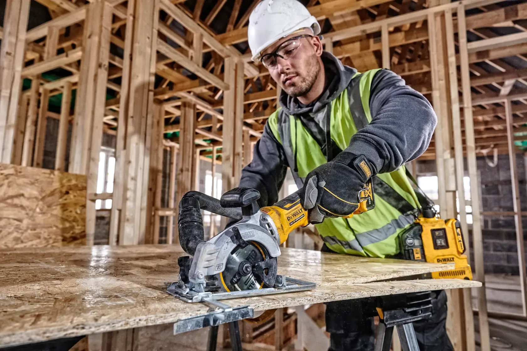 Operaio taglia legno con sega DEWALT in un edificio in costruzione, indossando dispositivi di sicurezza.