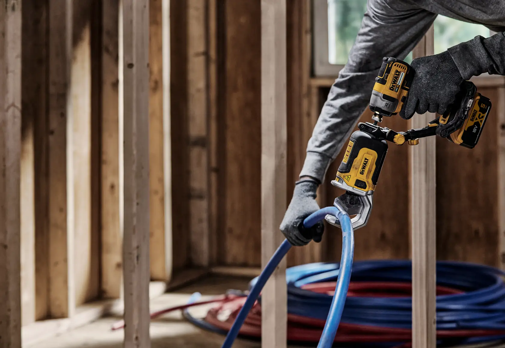 En person med hansker bruker et DEWALT DT20560 elektroverktøy til å kutte et blått plastrør i en trekonstruksjon.