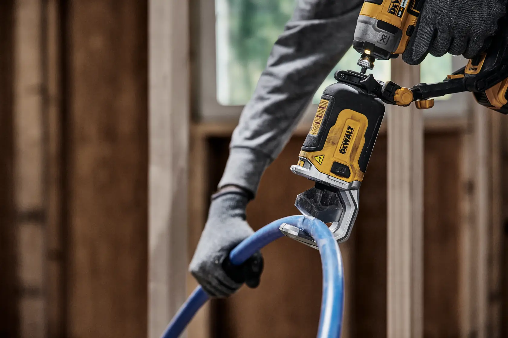 En person med hansker bruker et DEWALT DT20560 elektrisk verktøy til å kutte et blått plast­rør på en byggeplass.