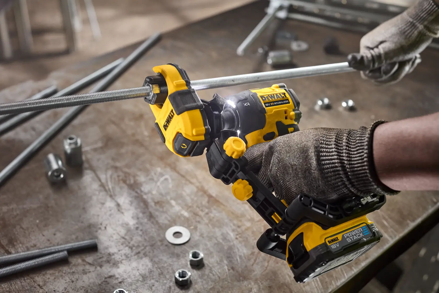 Gloved hands use a DEWALT power tool to cut a metal rod on a workshop table with nuts and washers.