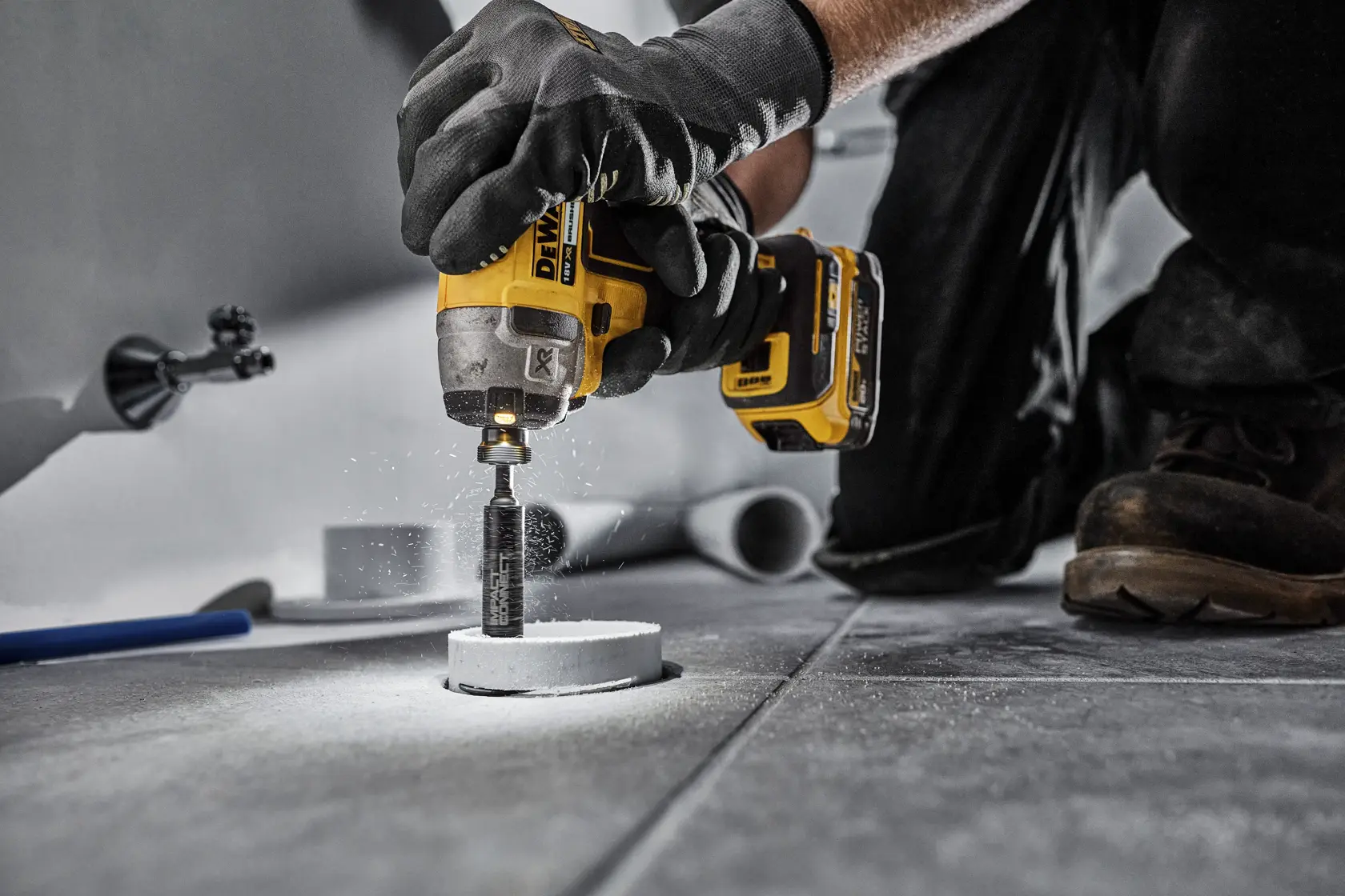 Person uses DEWALT drill tool with DT20563 accessory to cut through tile on a floor. Safety gloves are worn.