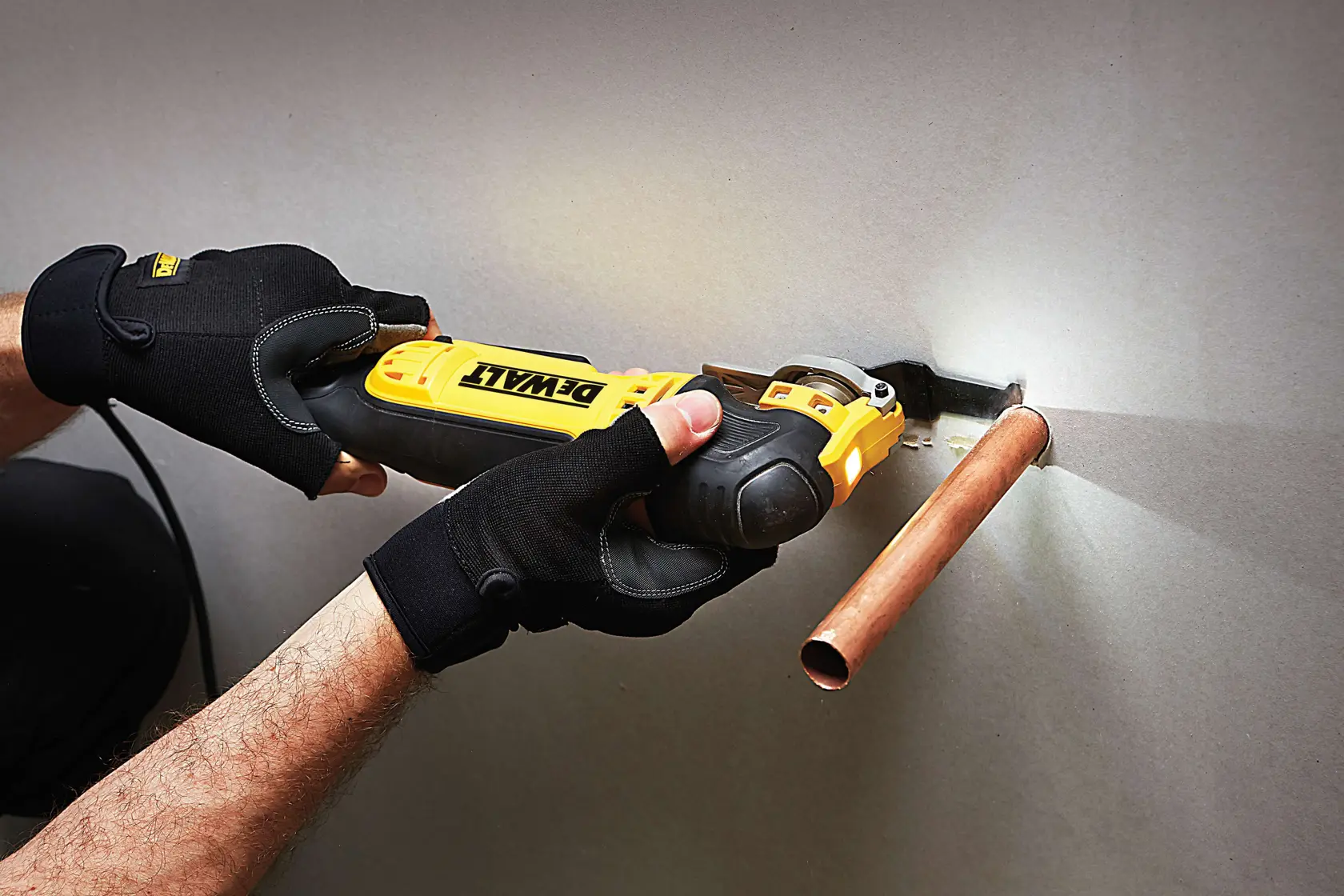 Person uses DEWALT tool to cut wall near copper pipe, wearing black gloves.