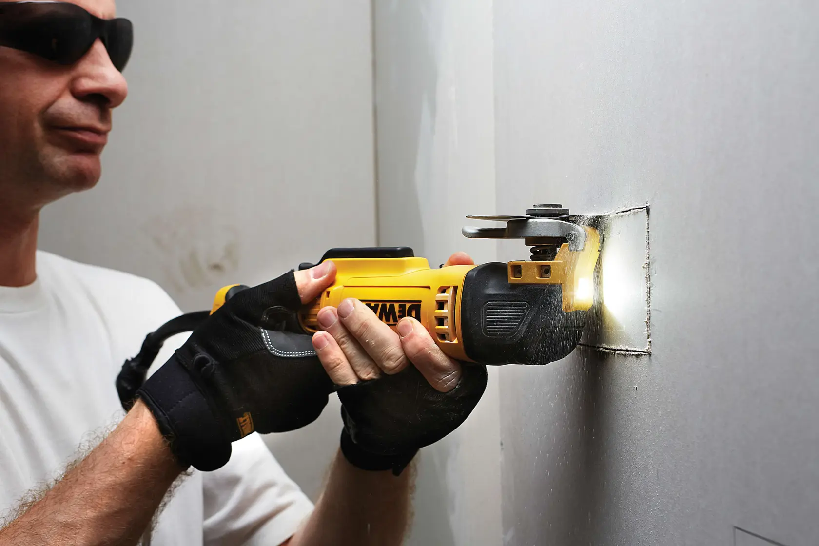 Person uses a DEWALT multitool to cut a square hole in drywall, wearing gloves for safety.