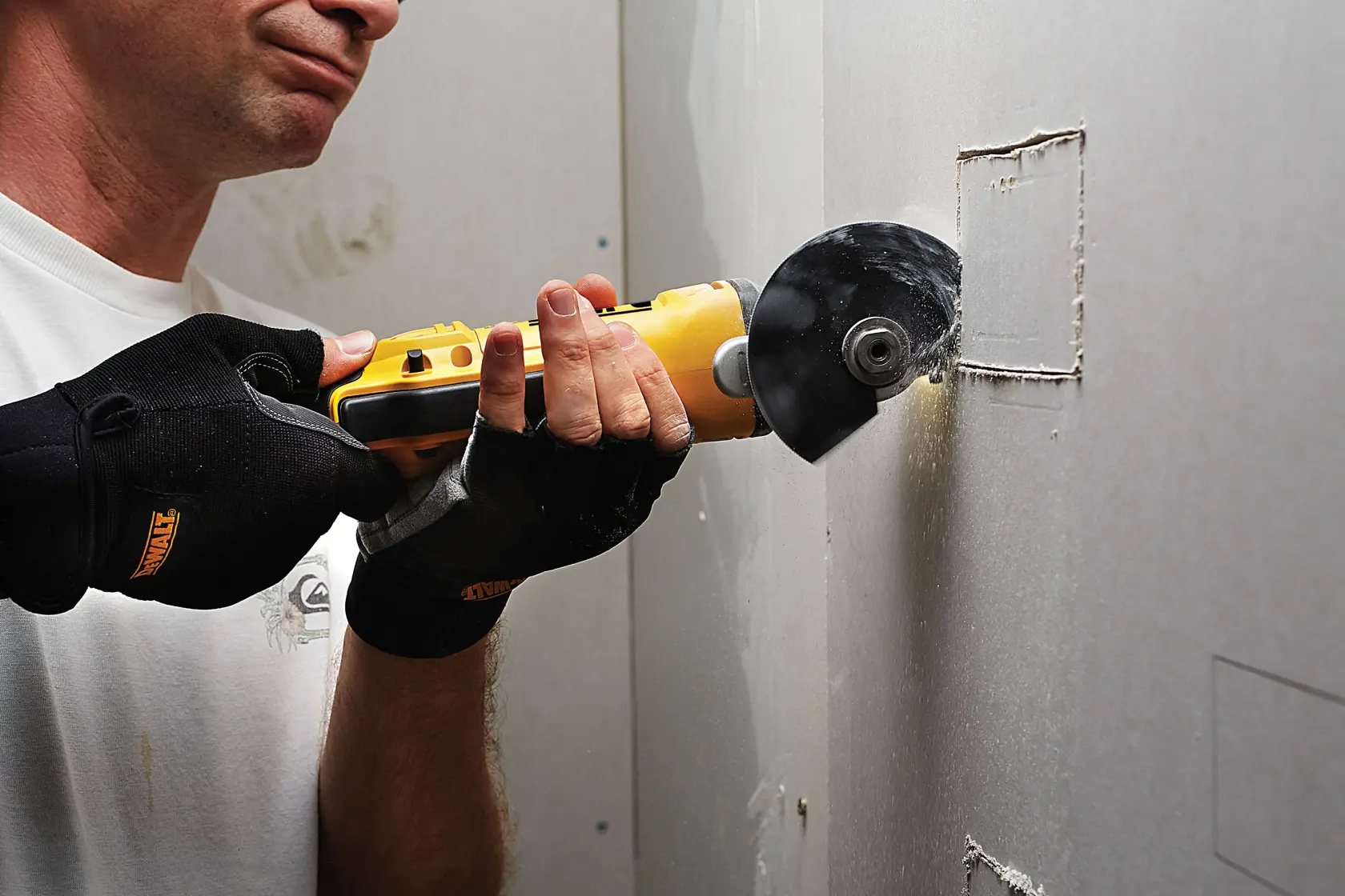 Person uses a DEWALT cutting tool to cut a square opening in drywall, wearing protective gloves.