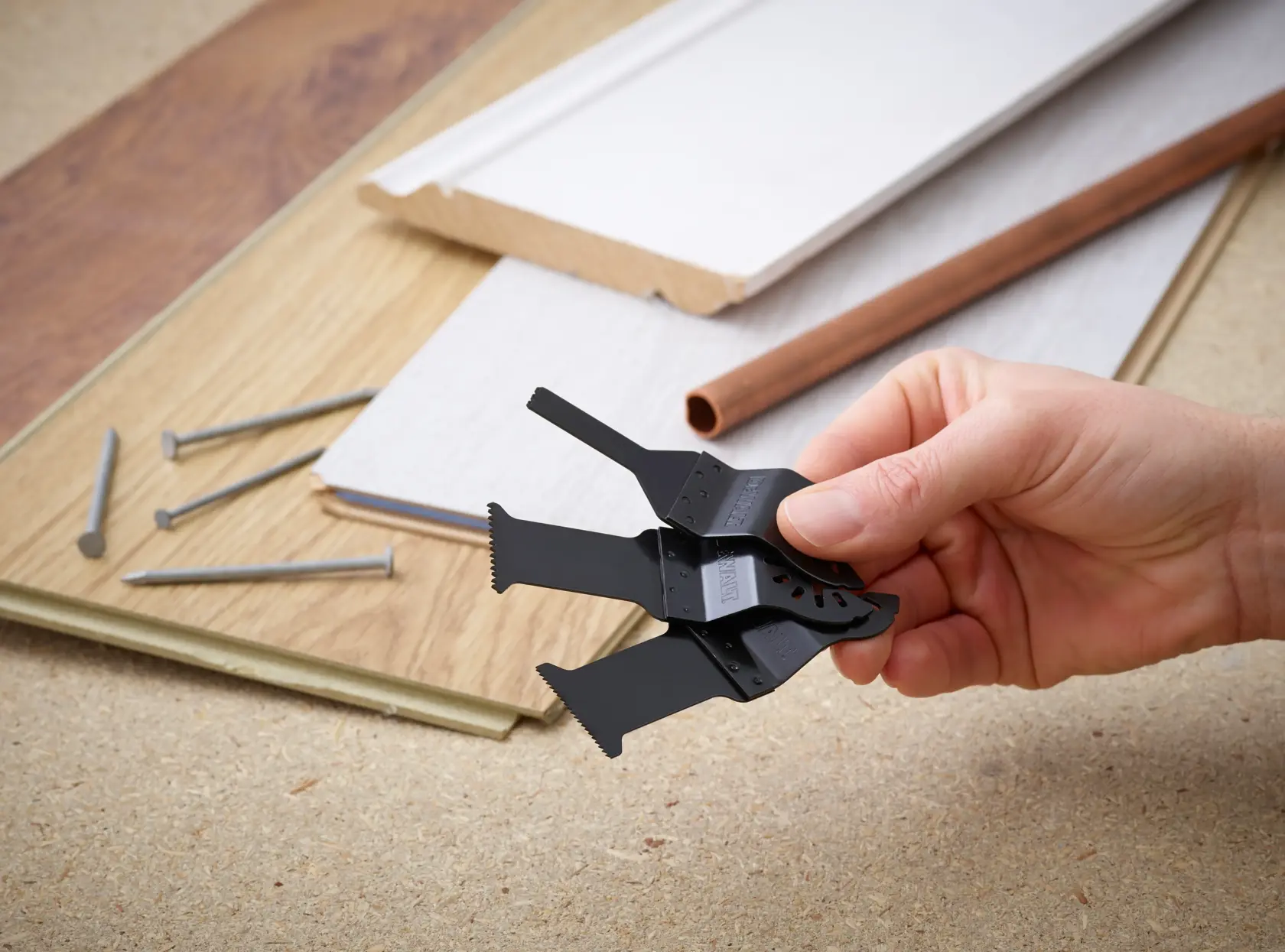 Hand holding DEWALT multitool blades, with nails, boards, and pipe on a workbench background.