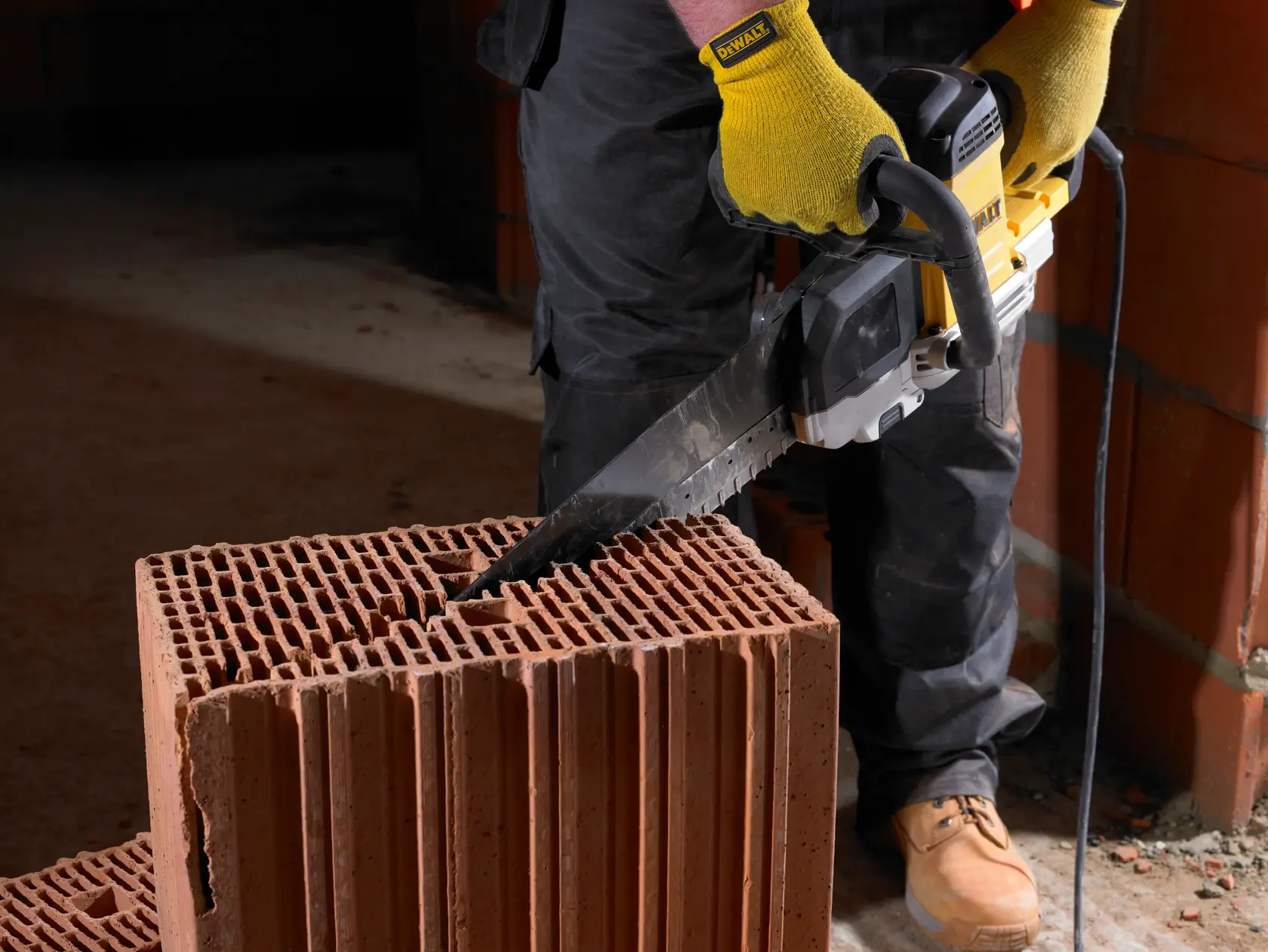 Person uses a DEWALT power saw to cut a hollow brick block indoors. Hands wear yellow gloves and boots are visible.