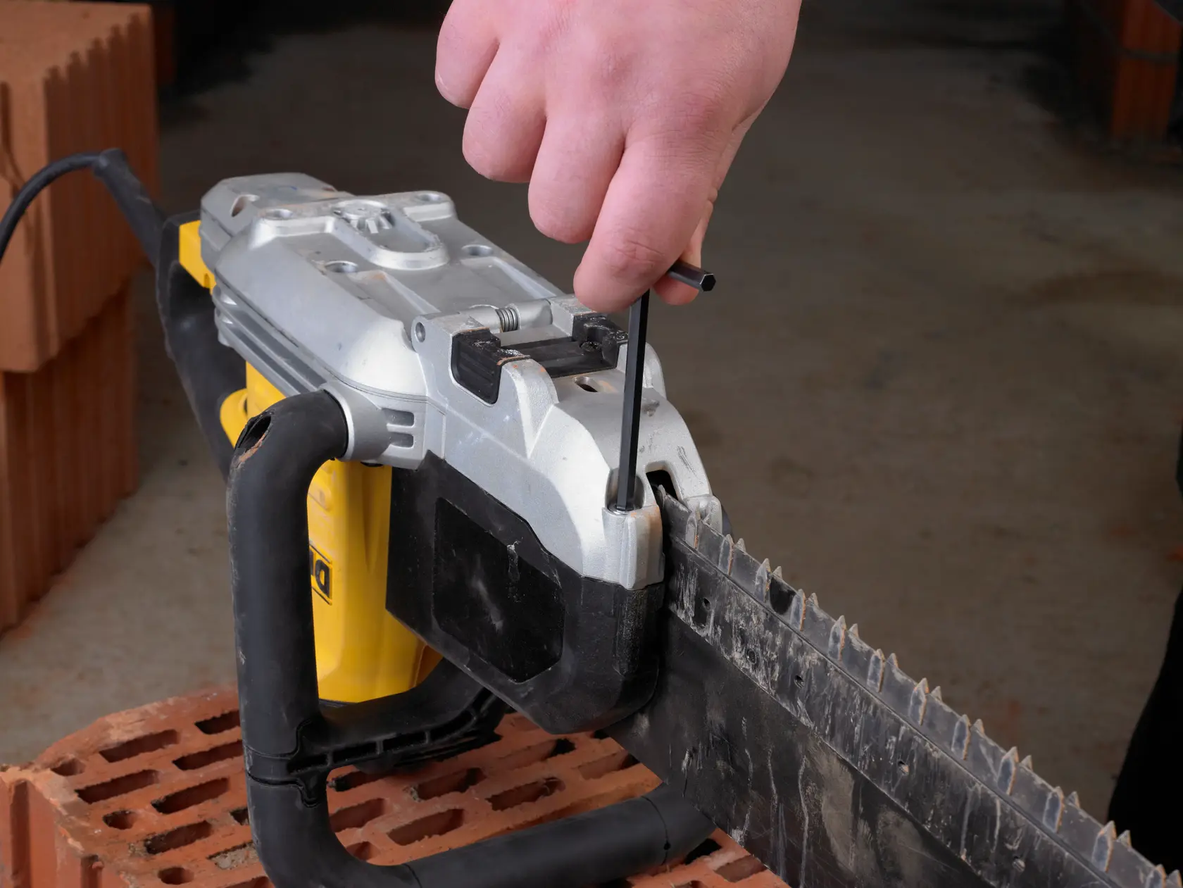 Hand uses a tool to adjust or tighten a DEWALT chainsaw blade, placed on a brick surface indoors.