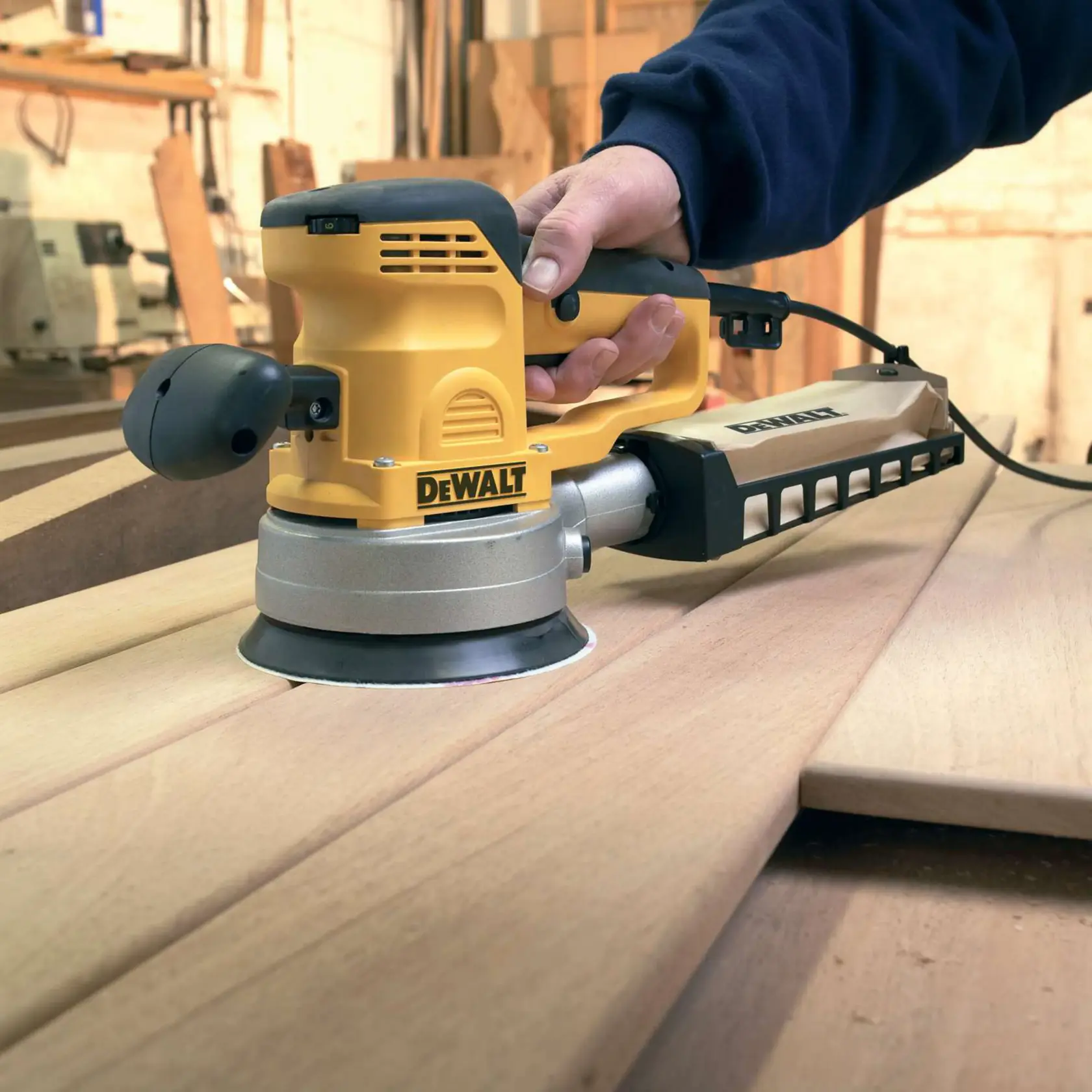 Levigatrice orbitale DEWALT usata su tavole di legno in una officina, tenuta da una persona con manica scura.