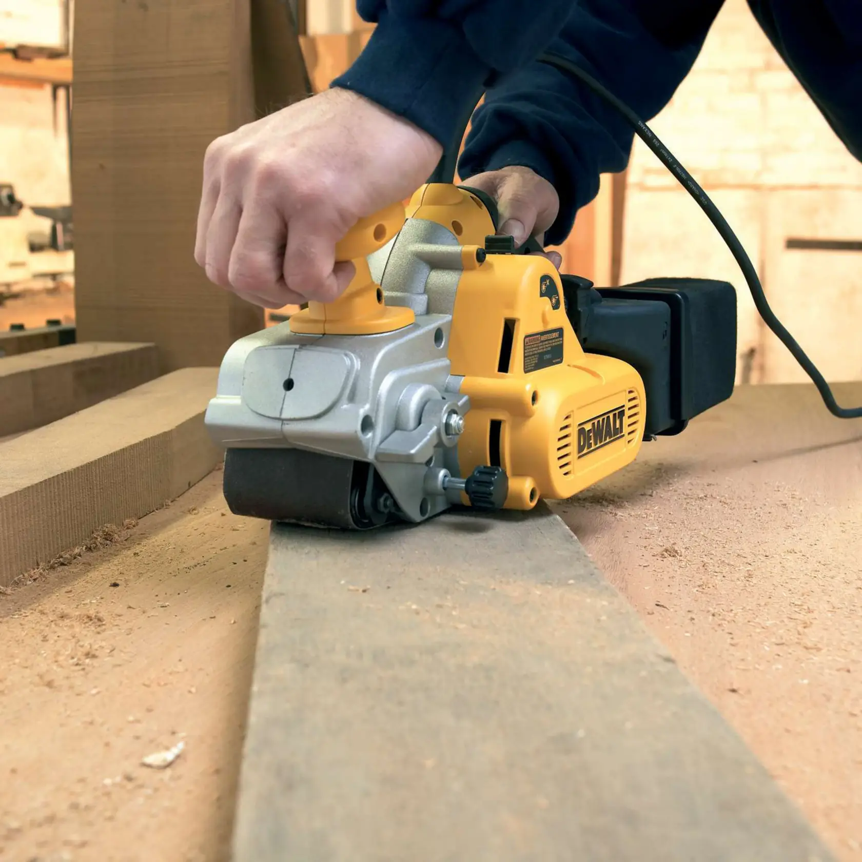 DEWALT belt sander in use, sanding a wooden plank in a workshop with sawdust visible on the surface.