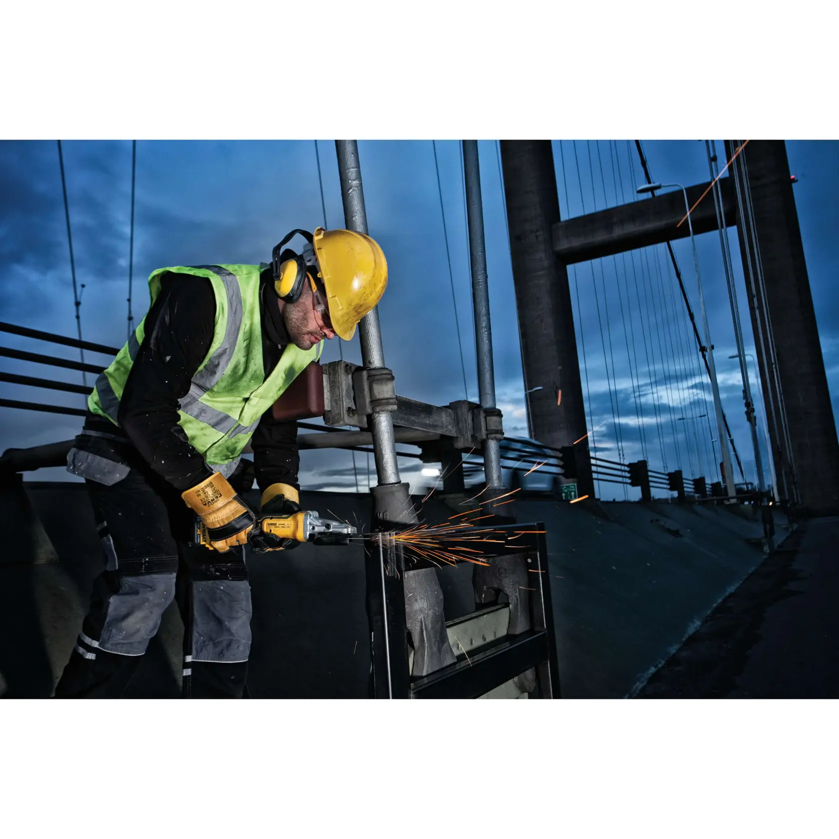 Worker in safety gear uses DEWALT tool on metal outdoors, sparks flying near a bridge at dusk.