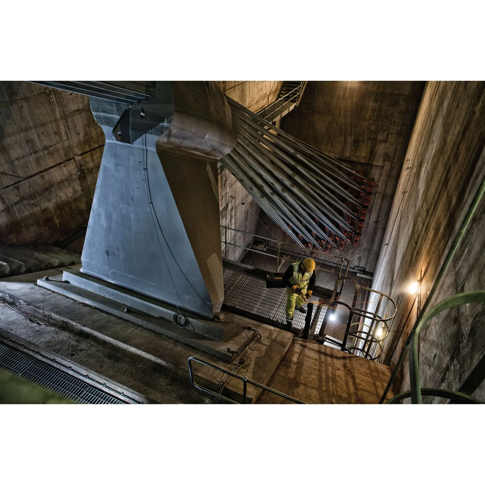 Worker in safety gear inspecting a large industrial structure with cables in a concrete facility with stairs.