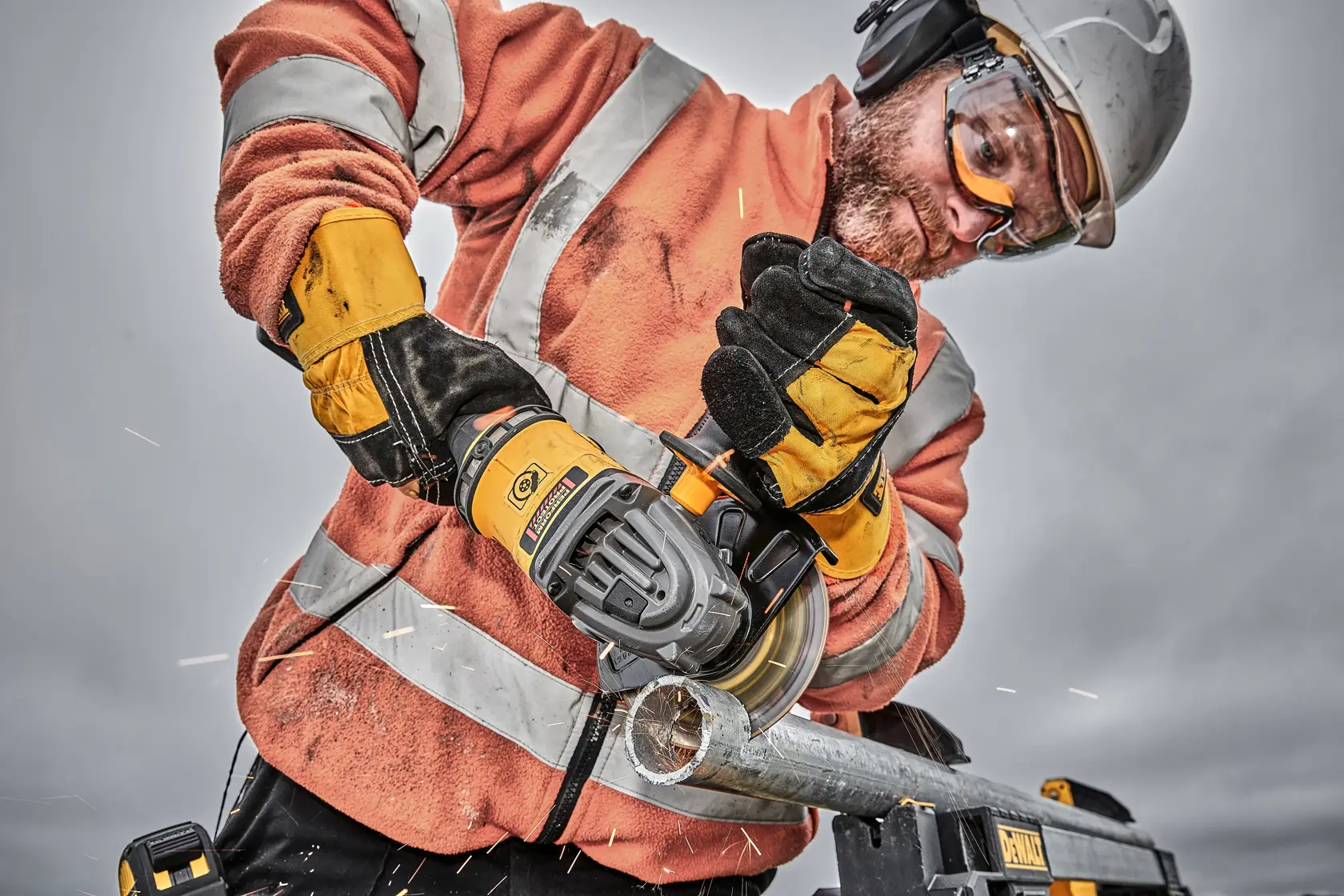 Worker using a DEWALT angle grinder to cut a metal pipe, wearing safety gear and gloves.