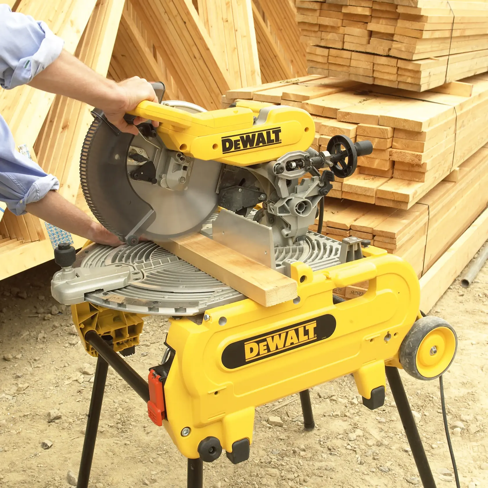 A DEWALT miter saw being used to cut a piece of wood, positioned on a yellow DEWALT stand. Stacks of wooden planks are visible in the background at an outdoor construction site.
