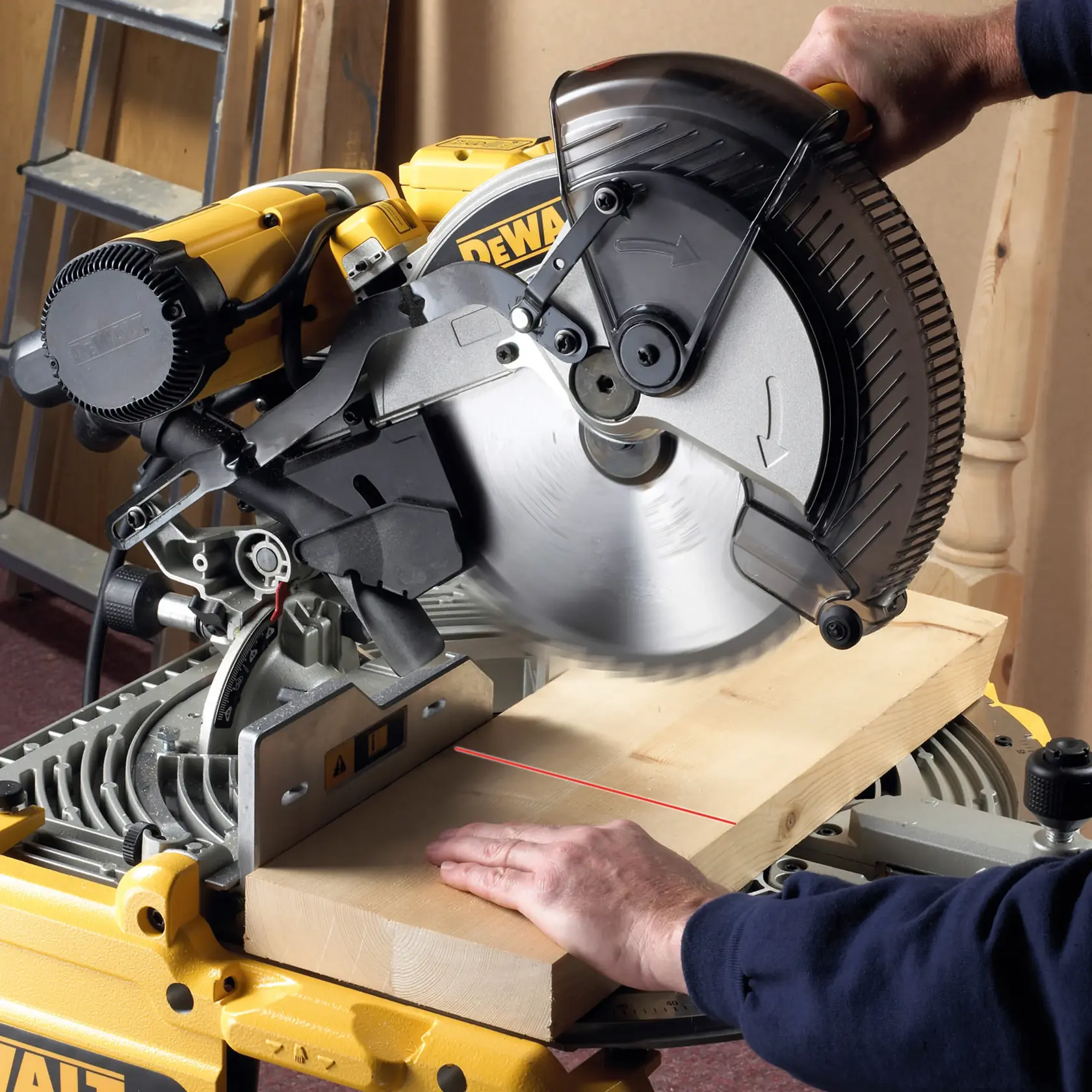 A DEWALT miter saw being used to cut a wooden plank. The saw blade is spinning and a person's hand is holding the plank steady on the saw table. The saw has a yellow and black design typical of DEWALT tools.