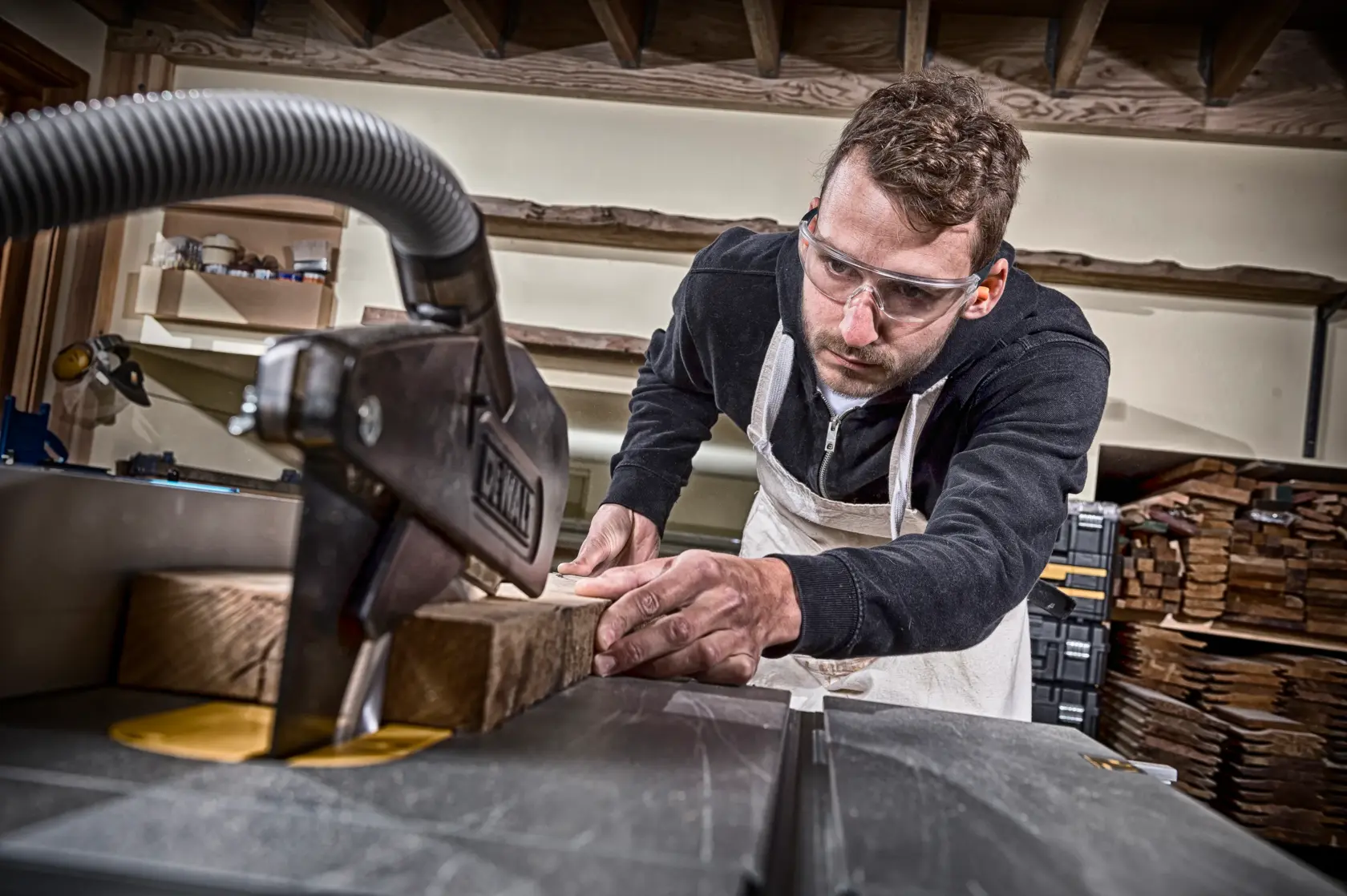 Person uses a DEWALT saw to cut wood in a workshop, wearing an apron. Stacks of wood are visible in the background.