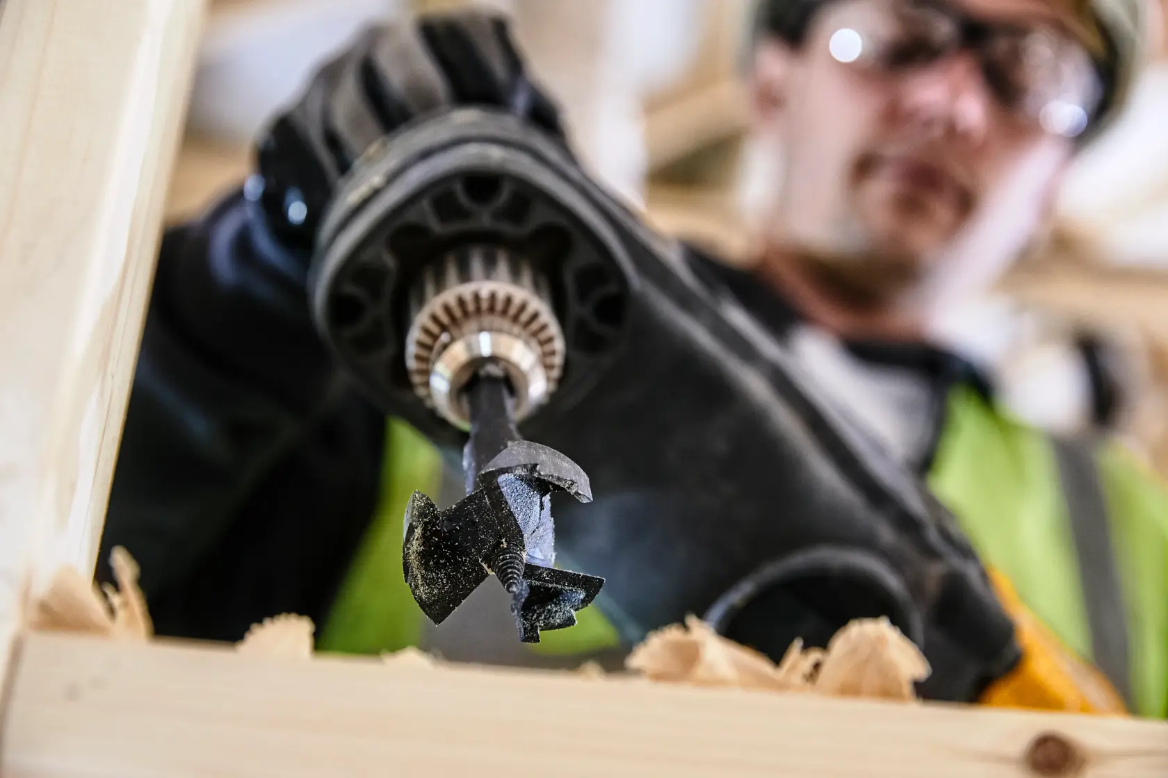 Primo piano di trapano DEWALT con punta DT4578 che fora legno, trucioli visibili, operaio sullo sfondo.