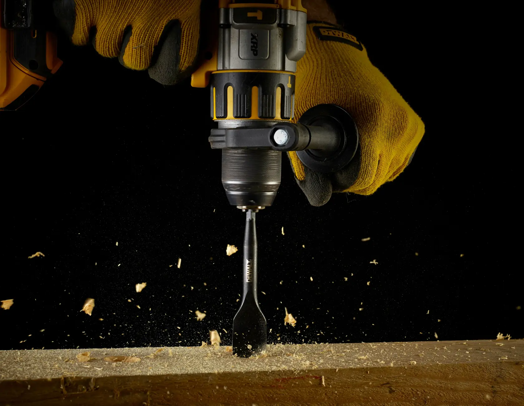Mano con guanto giallo fora il legno con utensile DEWALT, trucioli su sfondo nero.