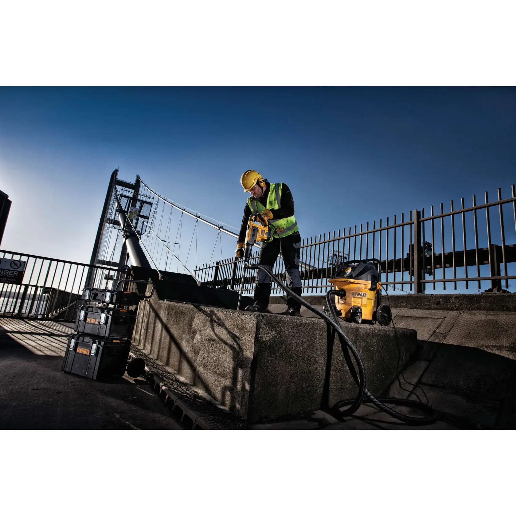 Worker uses DEWALT power tool and vacuum on concrete outdoors near a bridge, wearing safety gear and high-visibility vest.