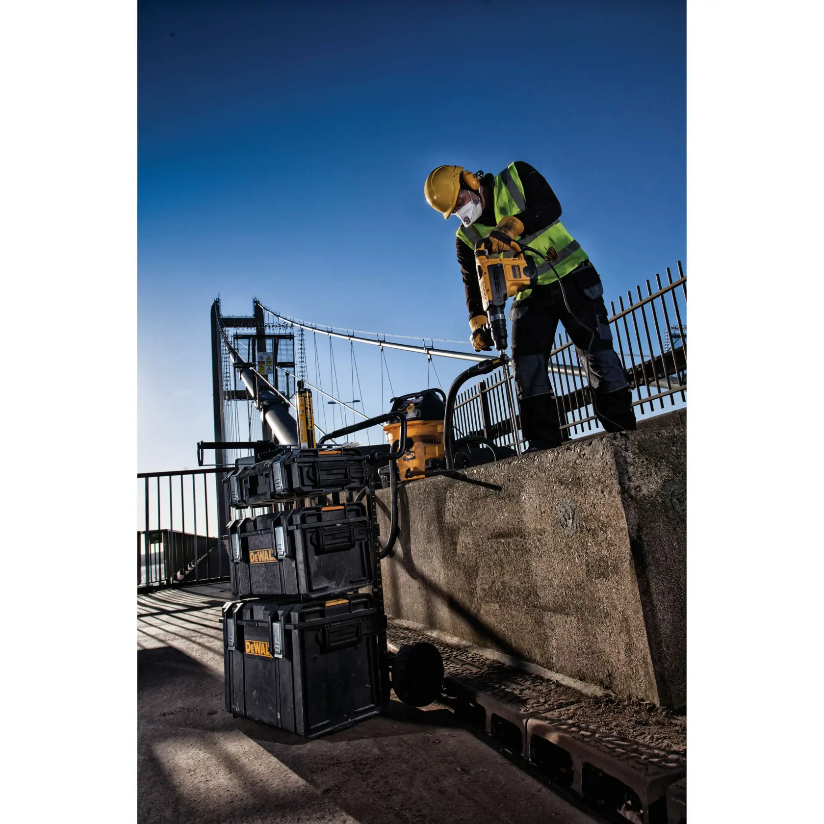 Worker uses DEWALT tools and storage boxes outdoors on a bridge construction site, wearing safety gear.
