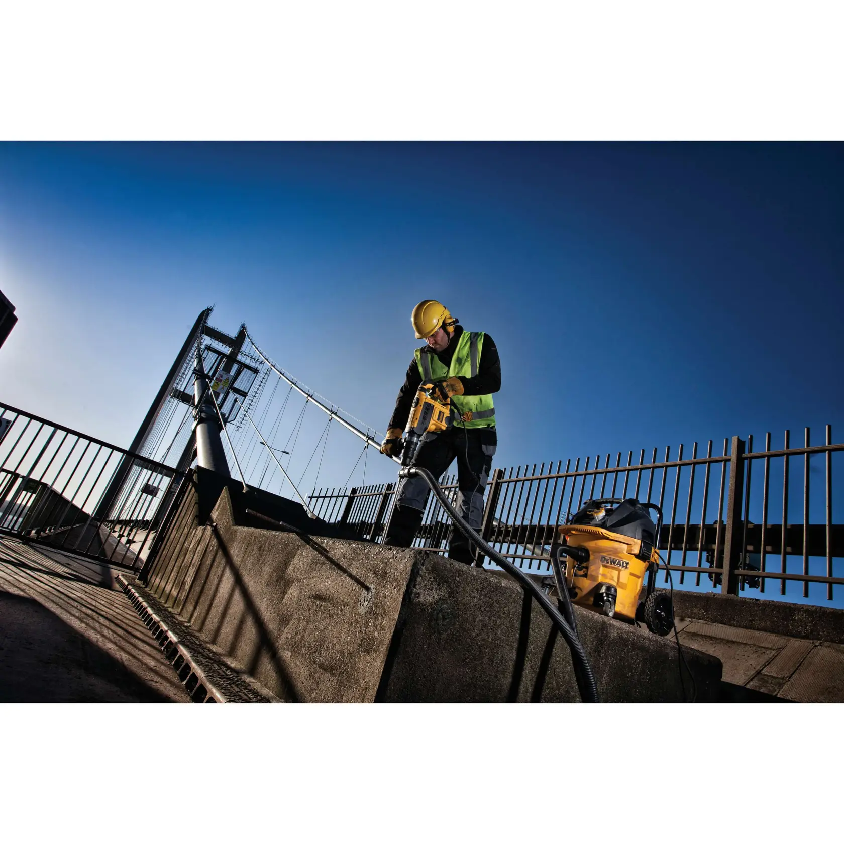 Worker uses DEWALT tool on concrete by bridge, wearing safety gear and helmet, with DEWALT equipment nearby.