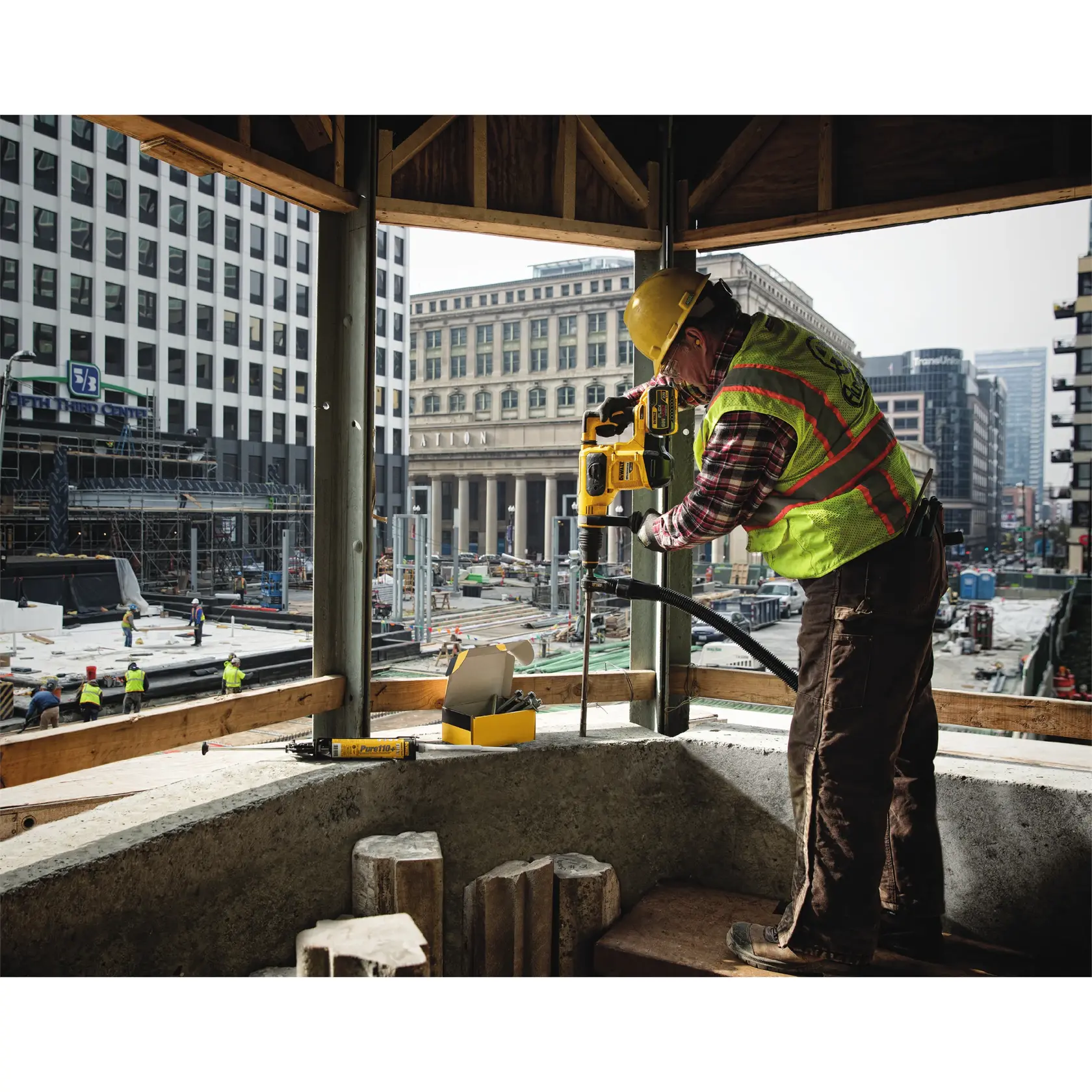 Worker using DEWALT DT60231 drill on construction site, wearing safety gear; city buildings in the background.