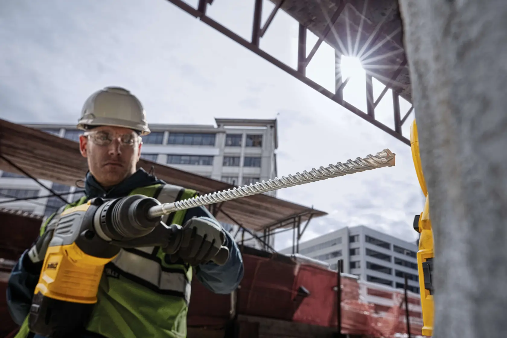 Worker uses DEWALT drill with DT60810 bit on concrete at a construction site; city buildings in background.