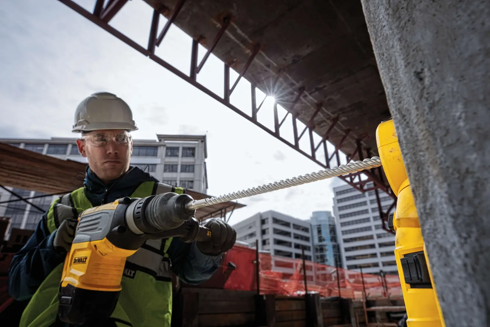 Worker uses DEWALT drill with DT60817 bit on a construction site, drilling into concrete near buildings.