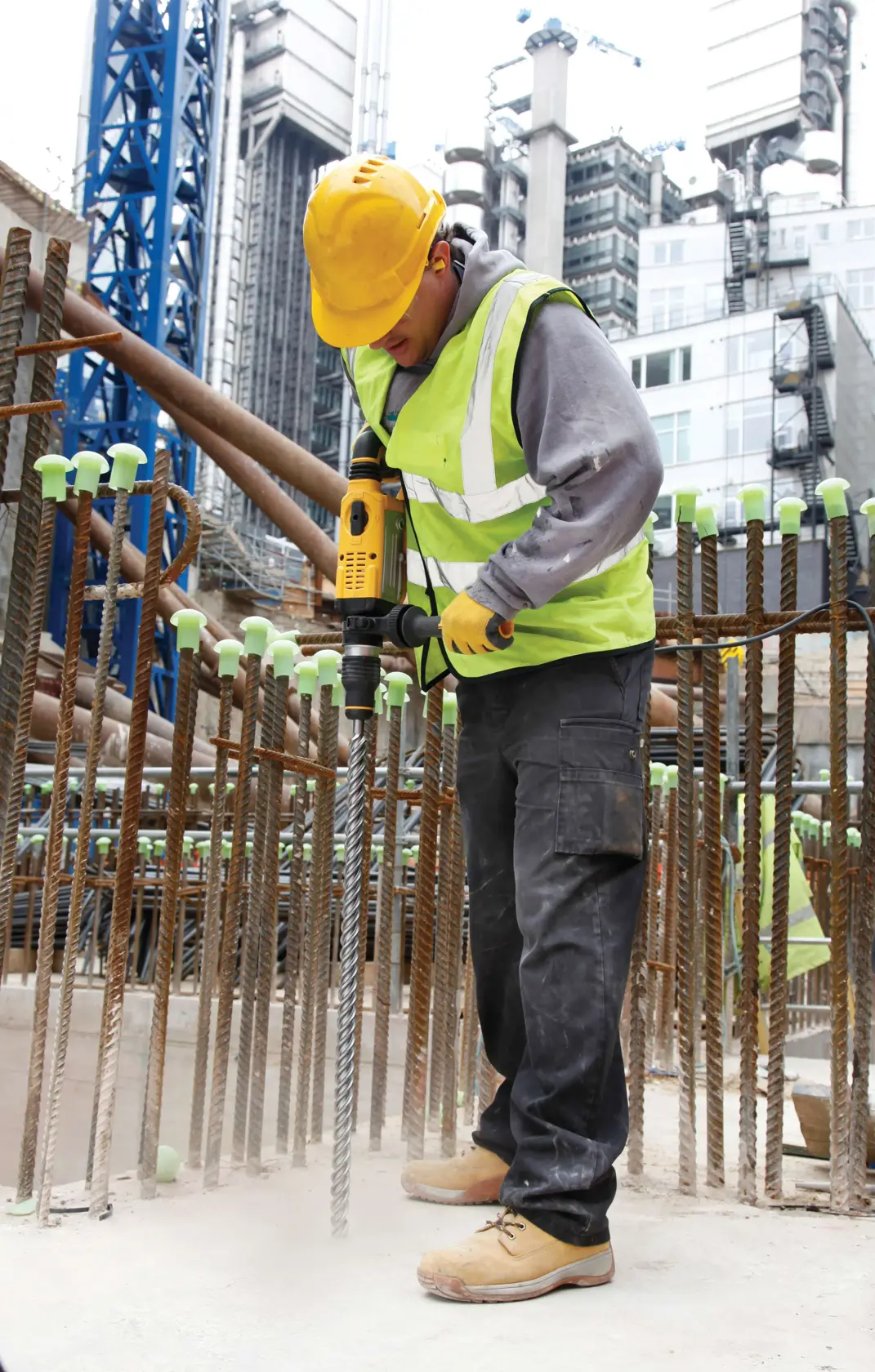 Worker uses a DEWALT hammer drill on a construction site, surrounded by rebar and wearing safety gear.