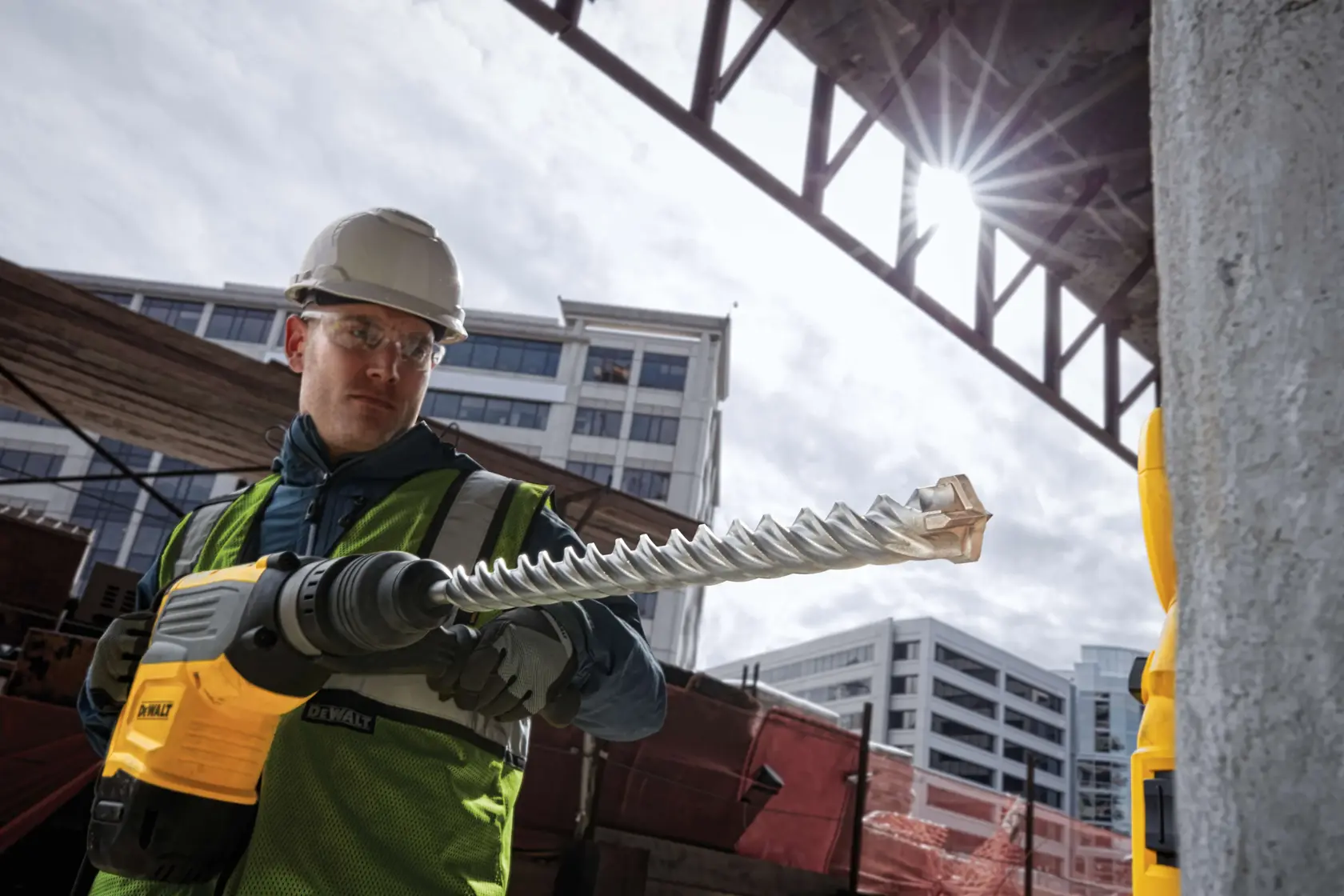 Worker uses a DEWALT hammer drill with a spiral bit on concrete at a construction site with modern buildings in background.