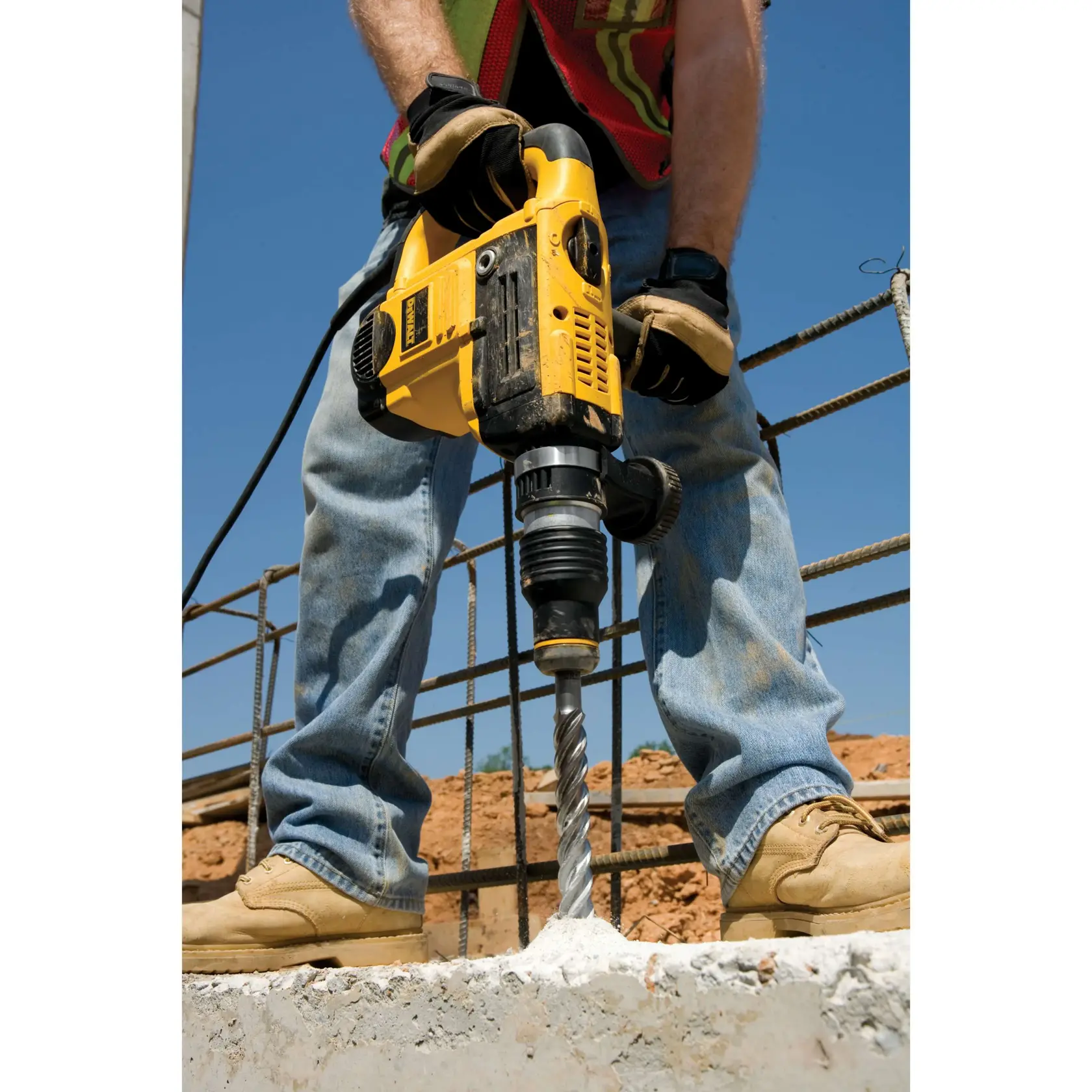 Worker uses a DEWALT power drill on concrete floor at a construction site with rebar and dust extraction.
