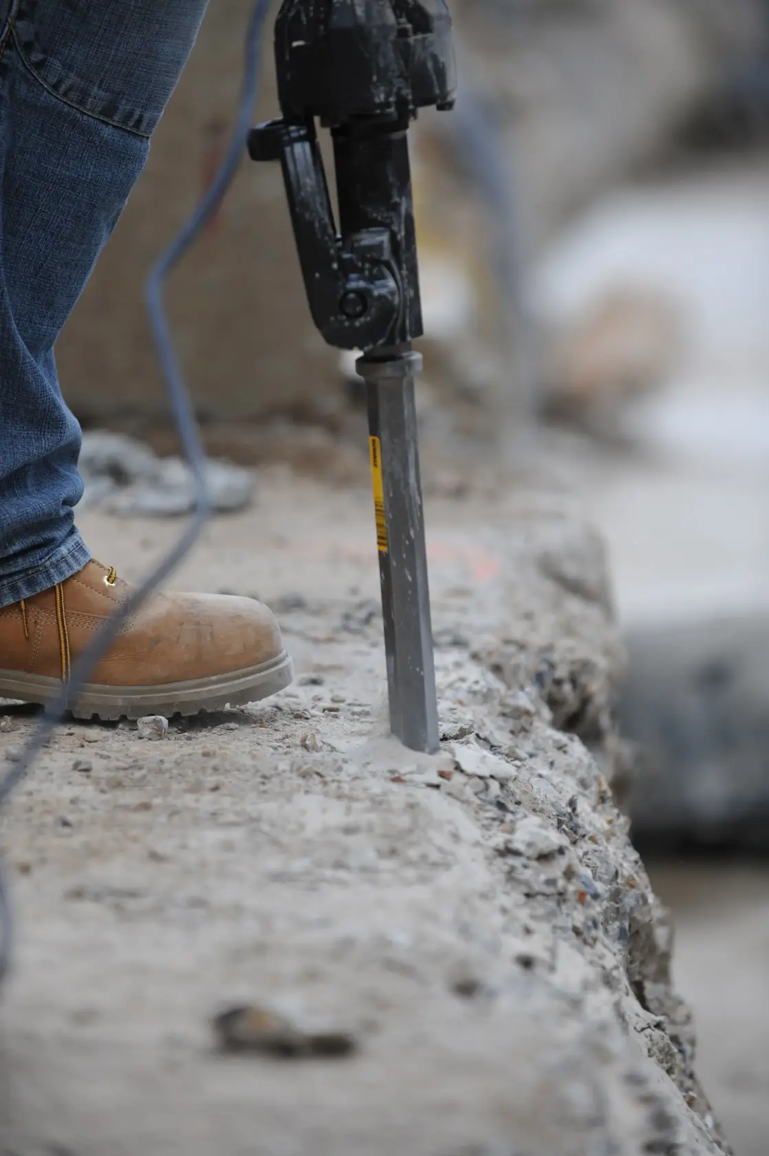 Worker uses a DEWALT chisel tool to break concrete on a construction site, wearing jeans and safety boots.