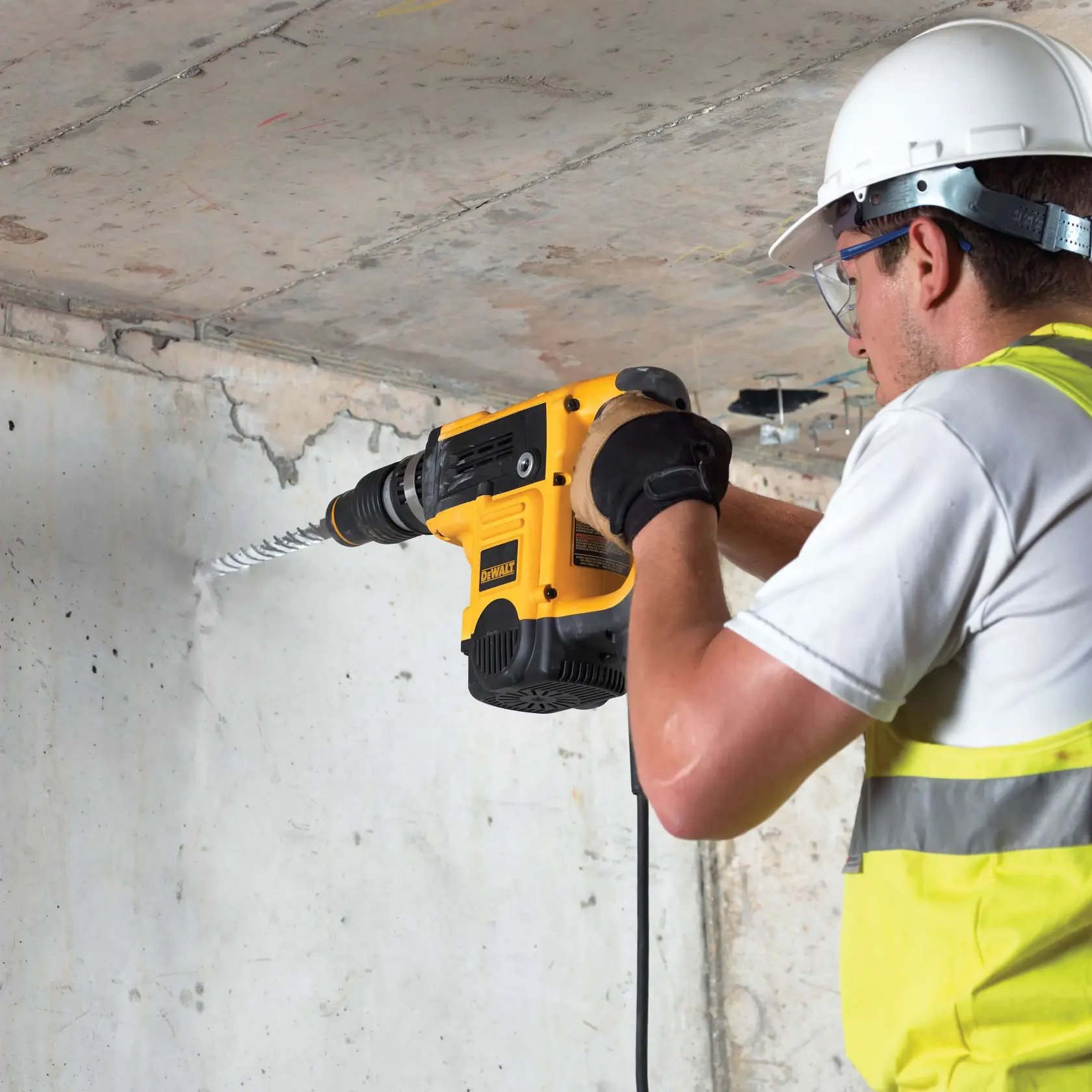 Worker drills into concrete wall using DEWALT DT9420 tool, wearing safety gear and high-visibility vest.