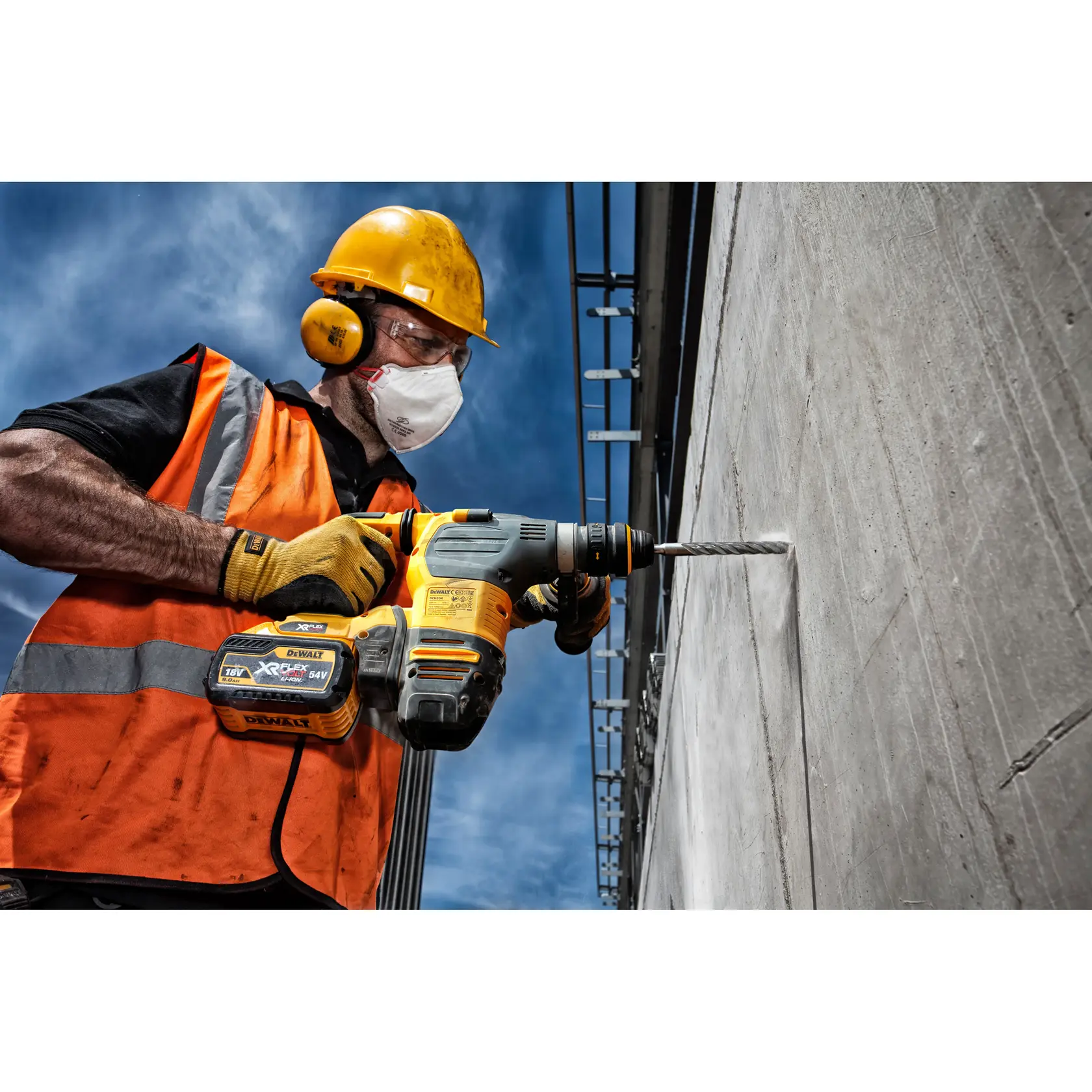 Worker uses DEWALT drill to bore a hole in a concrete wall, wearing safety gear and orange vest outdoors.