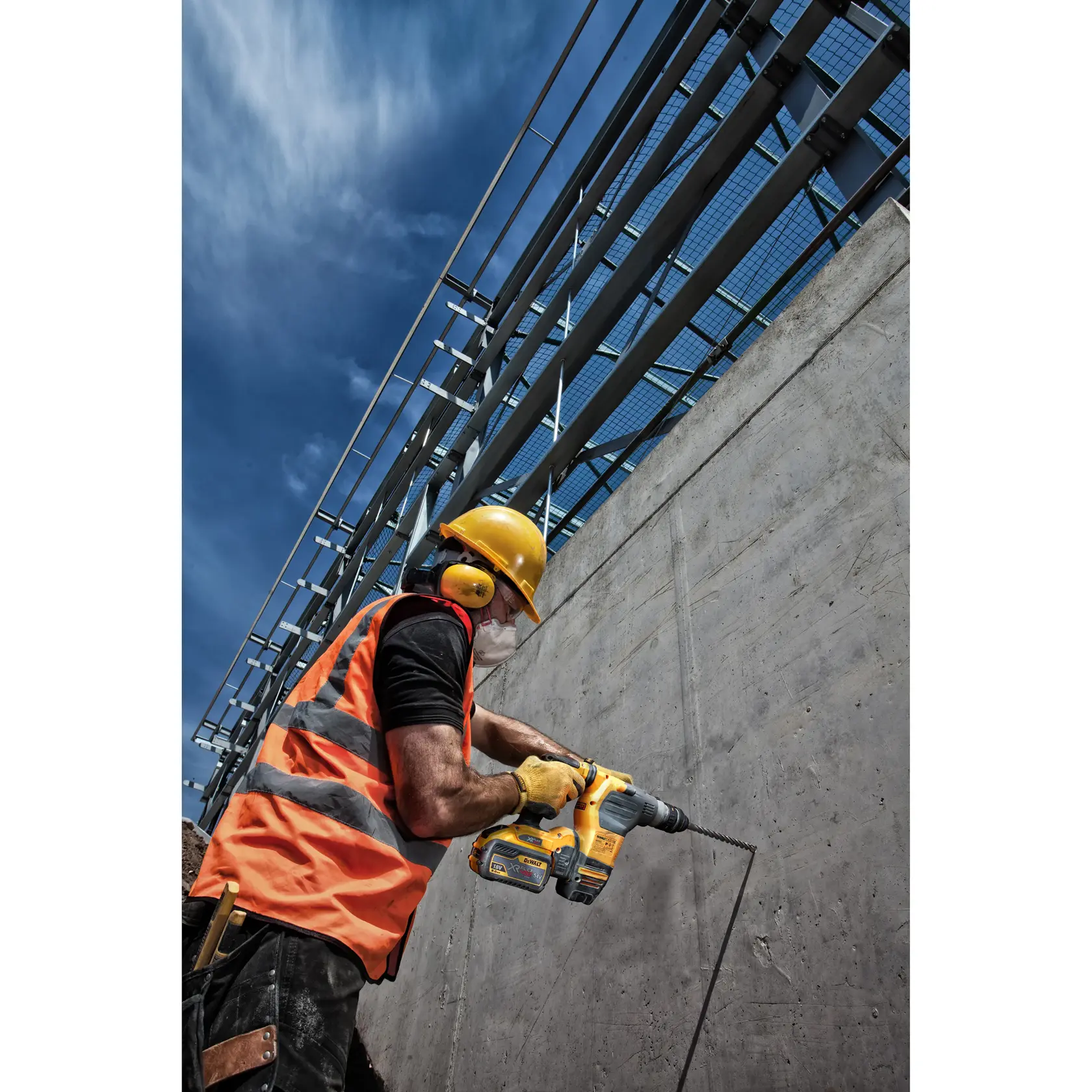 Worker uses a DEWALT drill to bore into a concrete wall outdoors, wearing safety gear and orange vest.