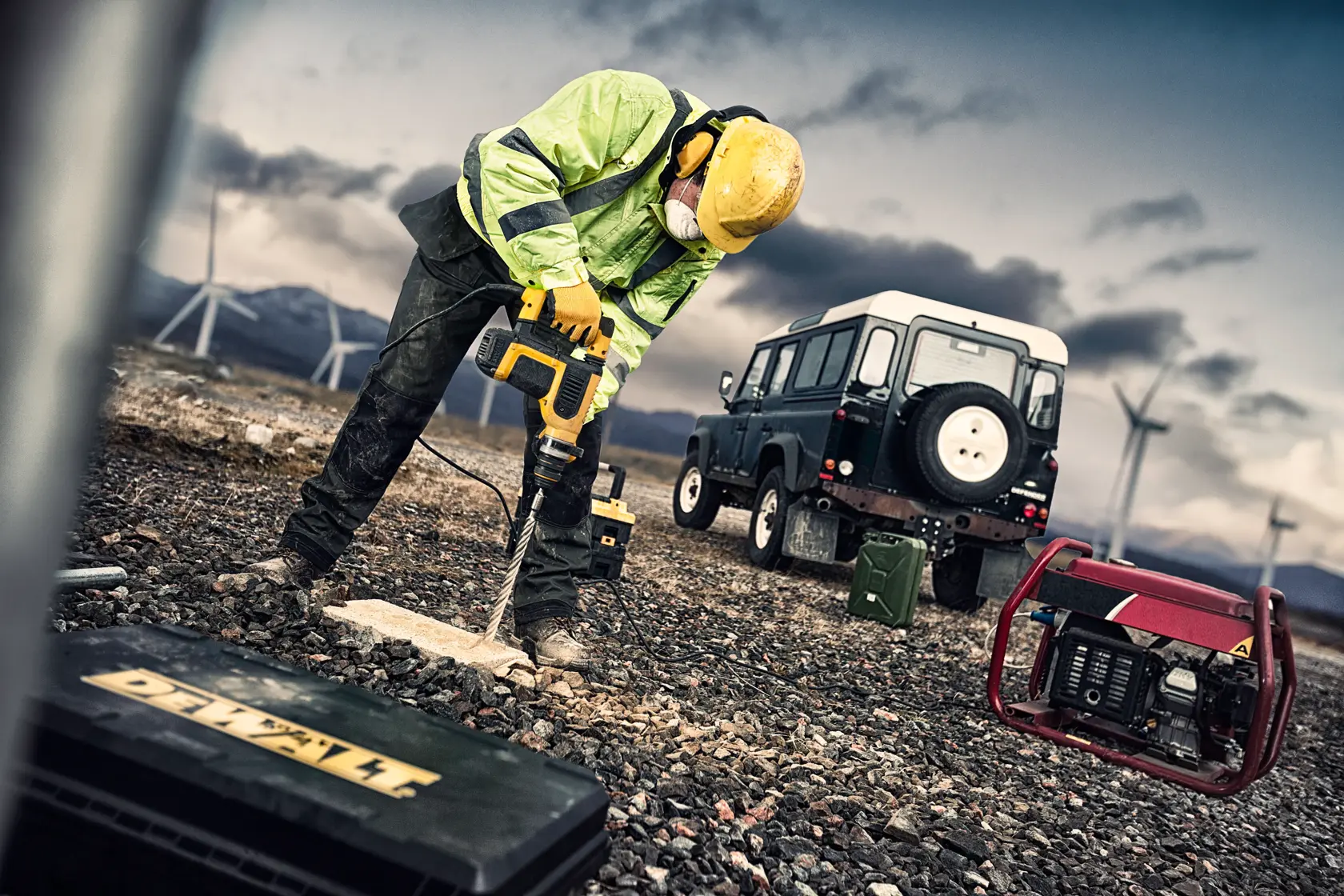 Trabajador con taladro DEWALT en suelo rocoso, vehículo, generador y aerogeneradores al fondo.