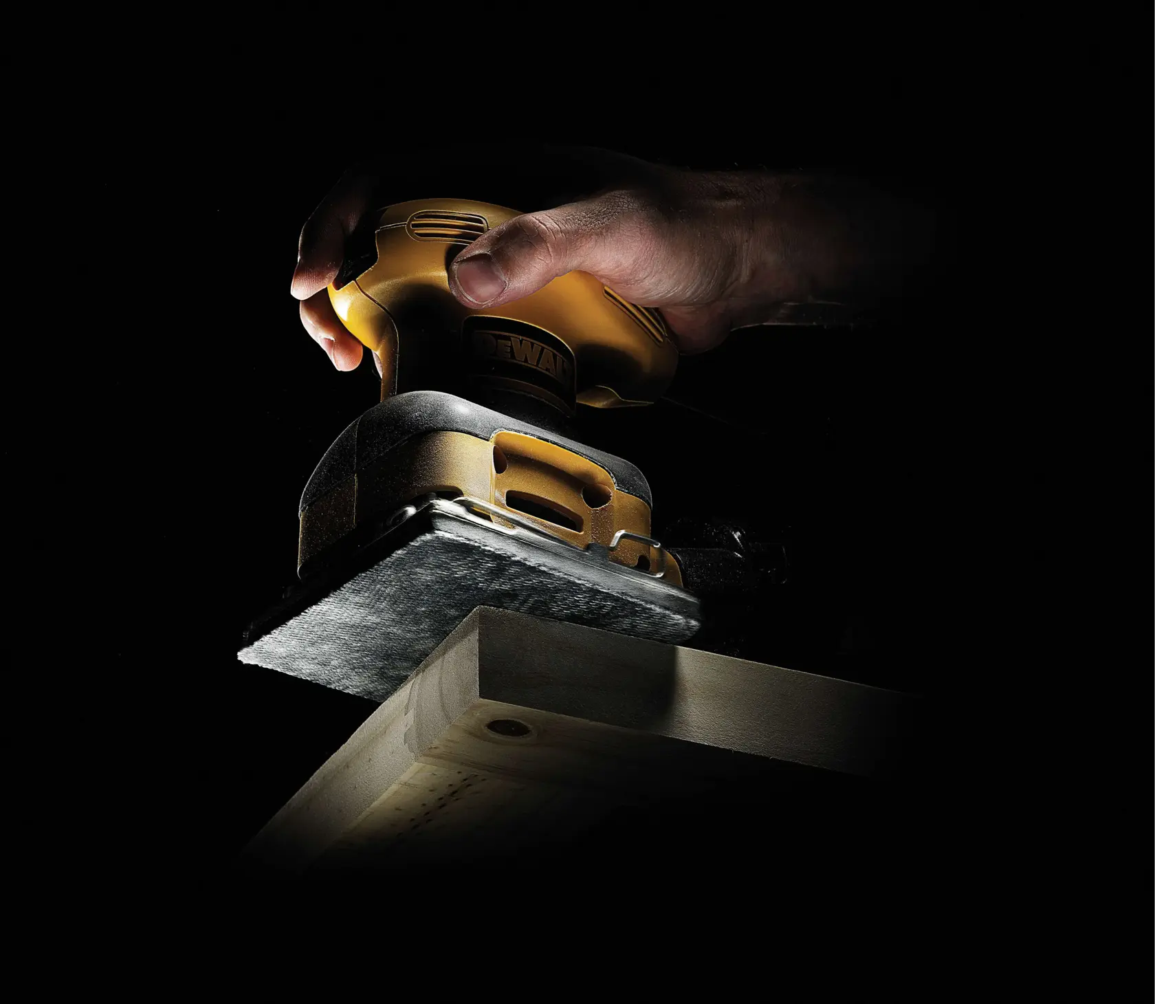 Hand using a DEWALT sander on a wooden plank in low light.