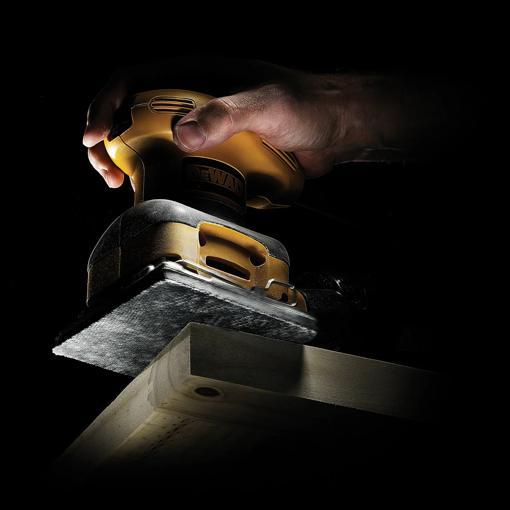 Hand holding a DEWALT sander, smoothing a wooden surface with the tool under focused lighting.