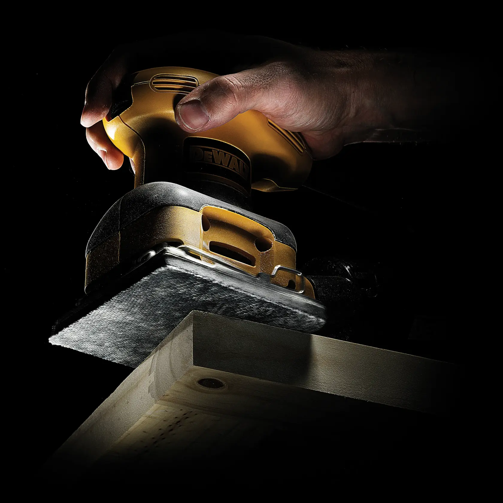 Hand using a DEWALT sander to smooth a wooden plank, close-up with dark background.